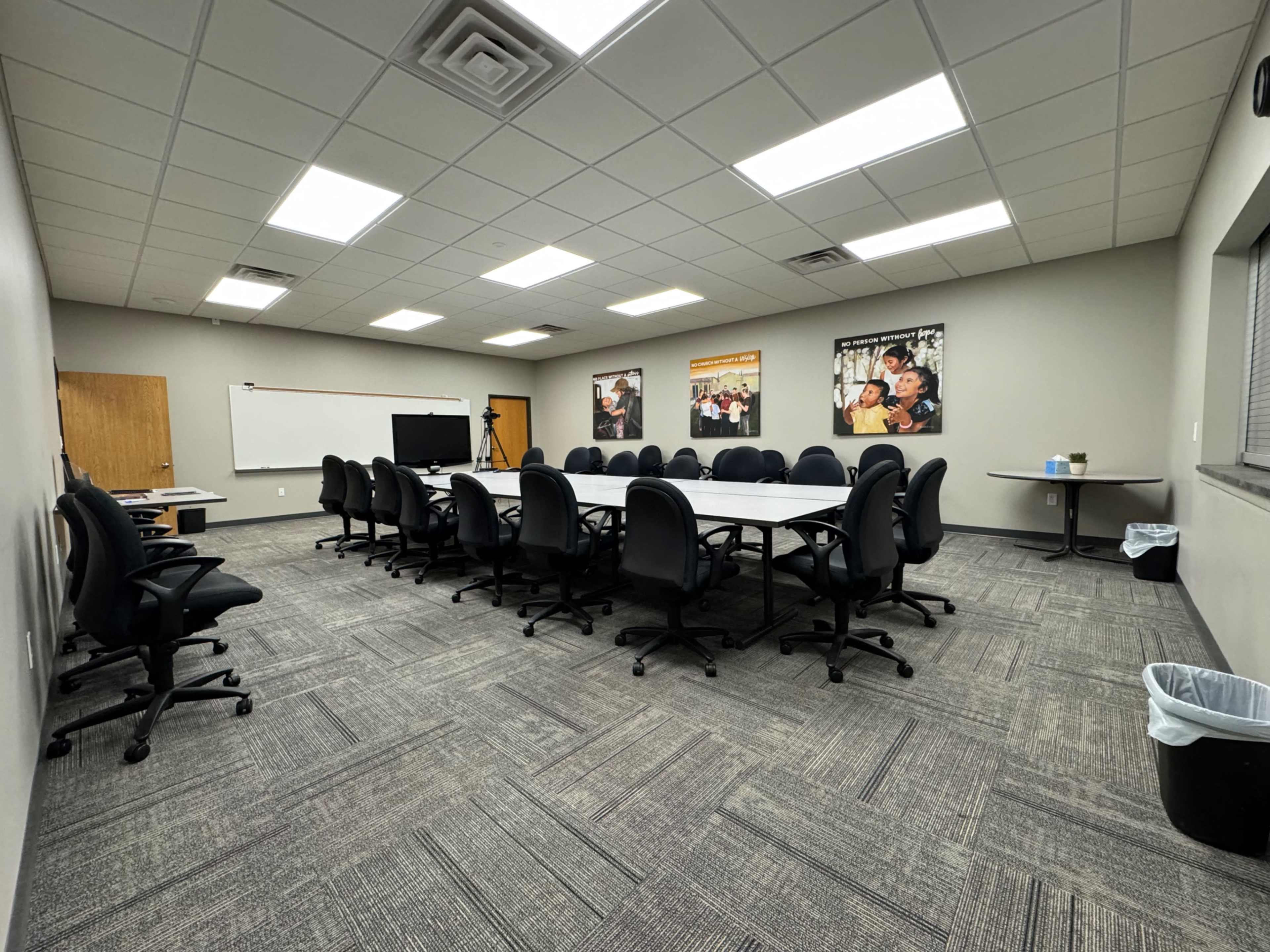 A large conference room features a rectangular table surrounded by black office chairs, with posters on the walls and a screen at one end.