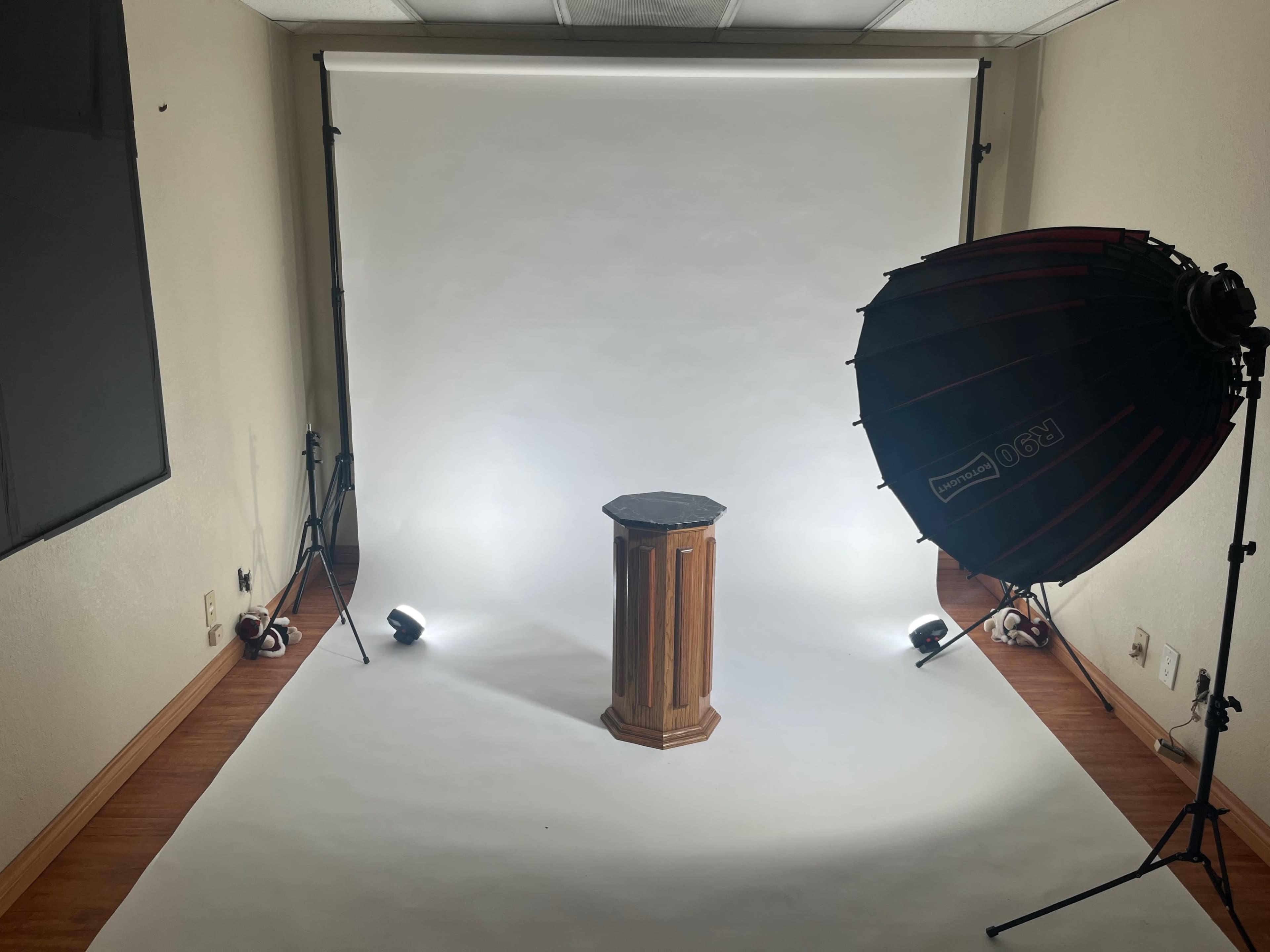 A photography studio with a wooden pedestal in the center, surrounded by lighting equipment and a white backdrop.