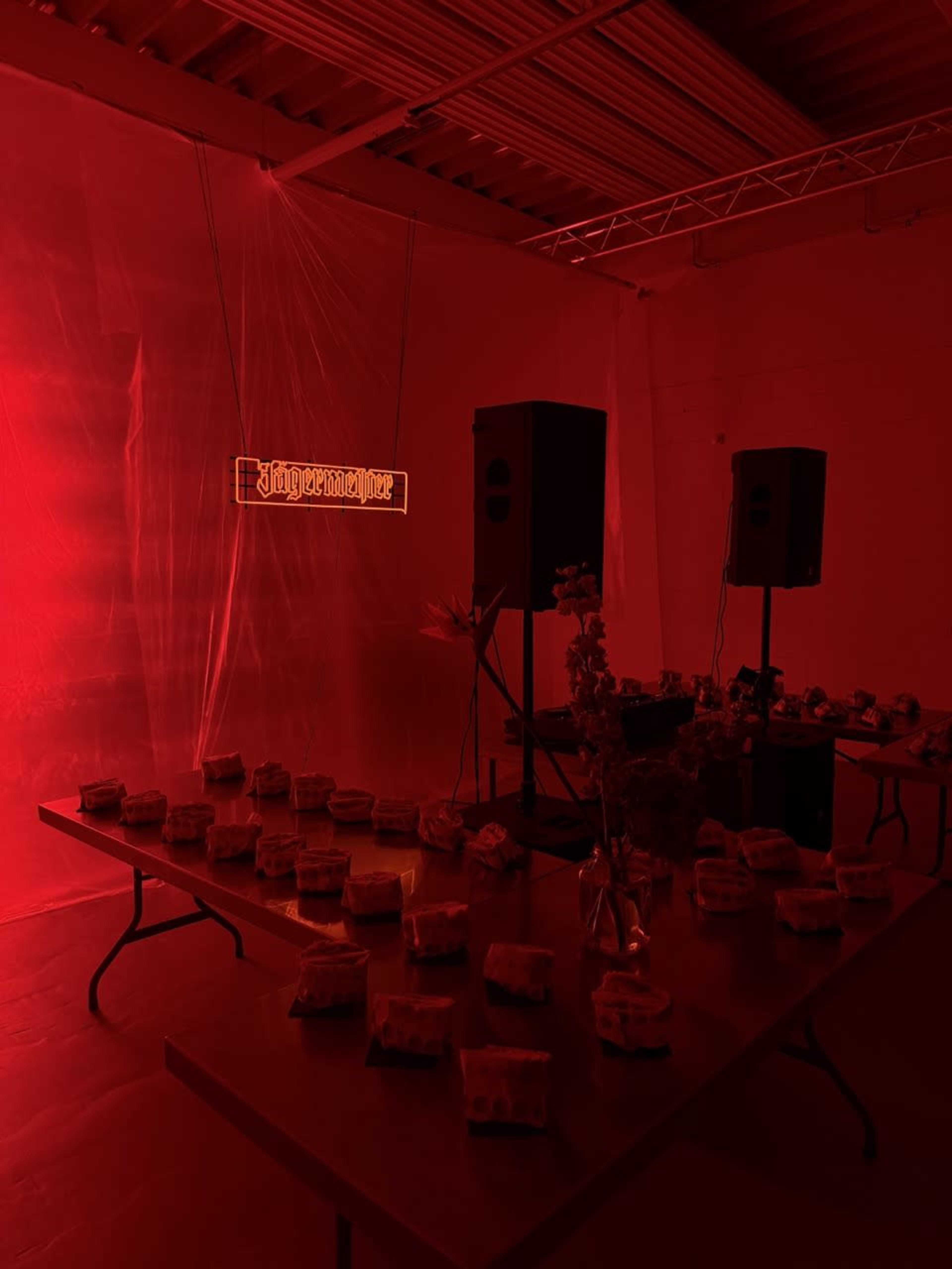 The image shows a dimly lit room with red lighting, featuring a table arranged with packages and two speakers, along with a neon sign that reads "Judgment."