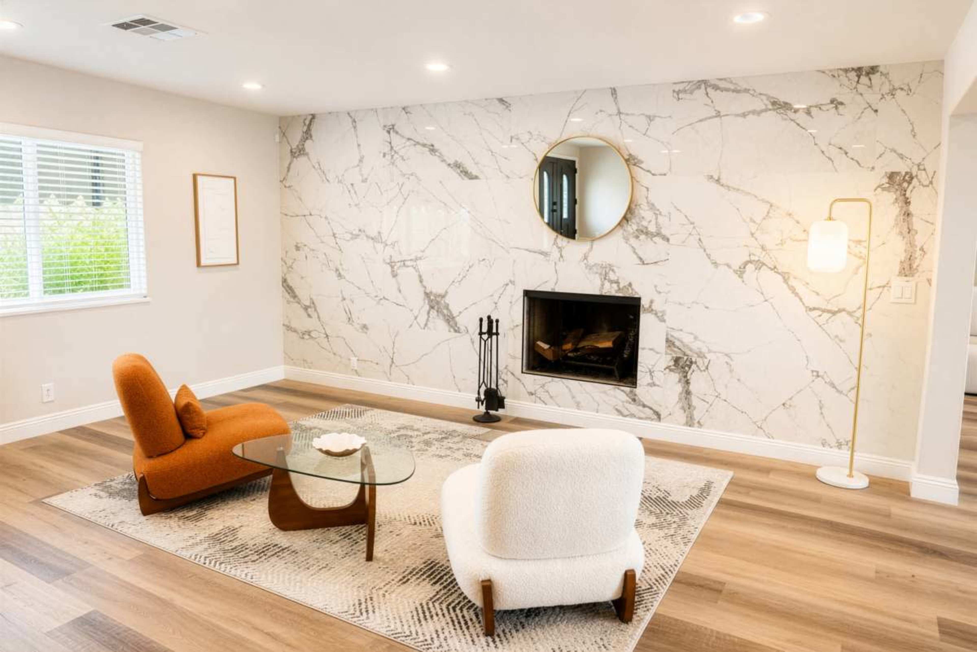 The image depicts a modern living room featuring two upholstered chairs, a glass coffee table, a fireplace, and a marble accent wall.