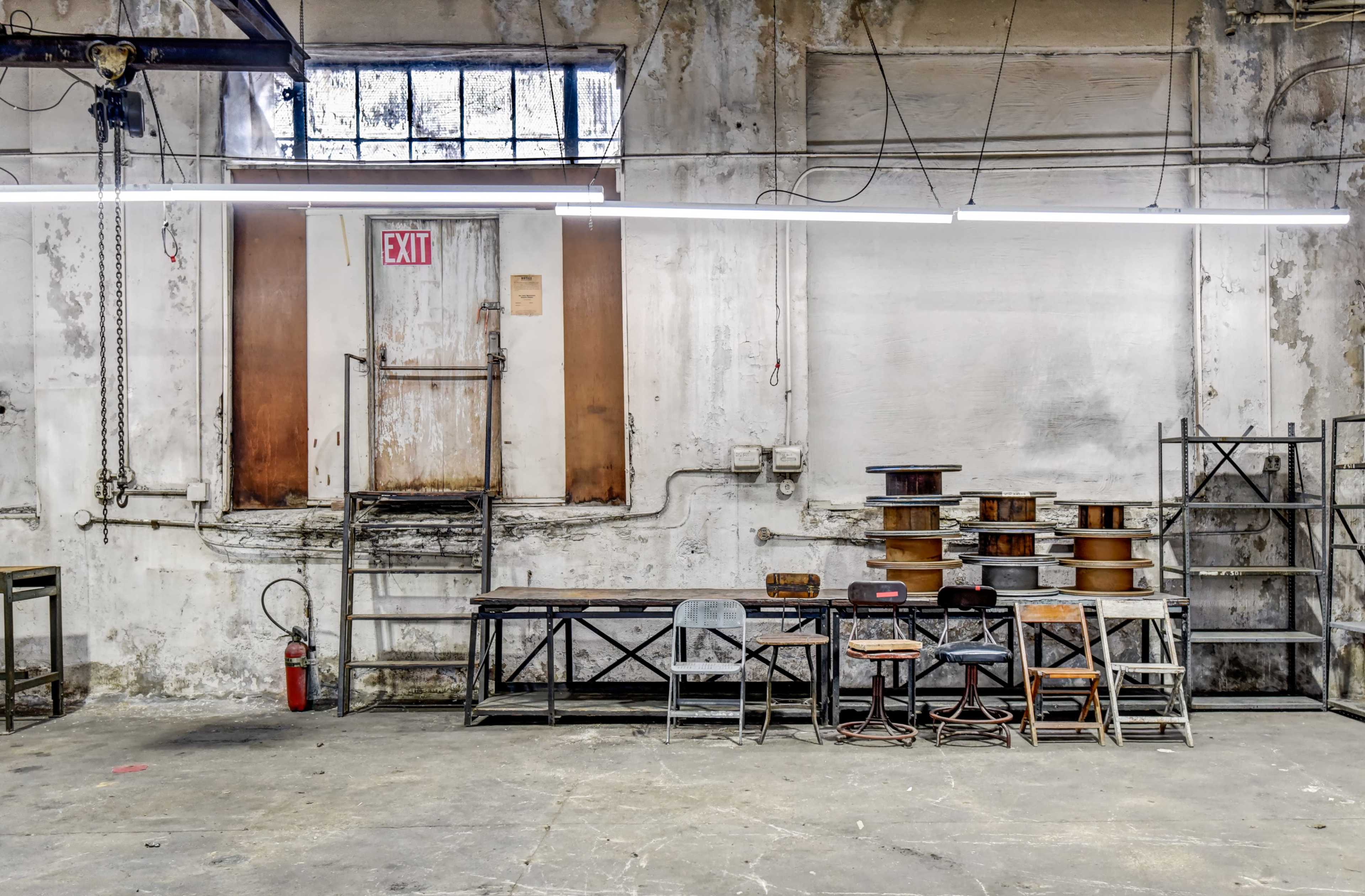 A spacious, empty industrial workshop with various metal and wooden furniture, including tables and stools, along one side of a weathered concrete wall.