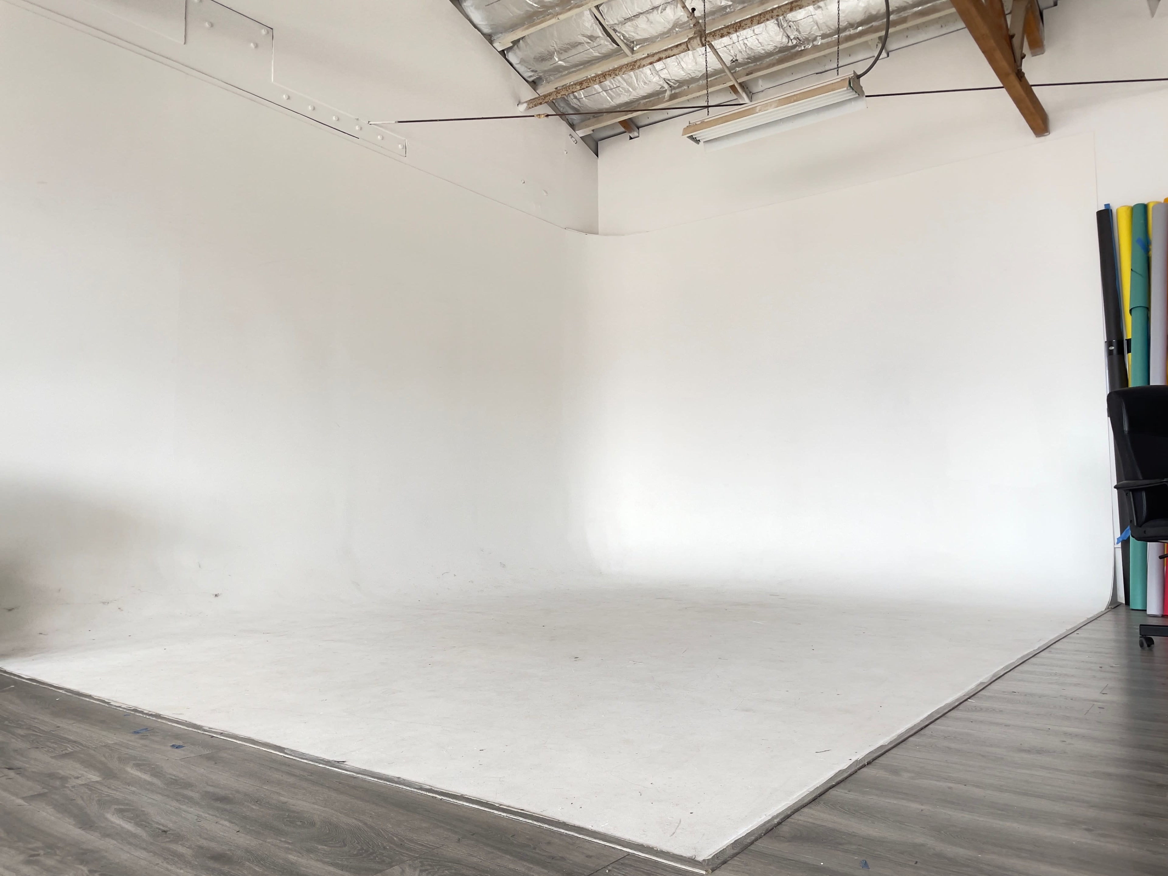 A spacious, empty studio with a white wall, wooden beams, and a black chair in one corner.