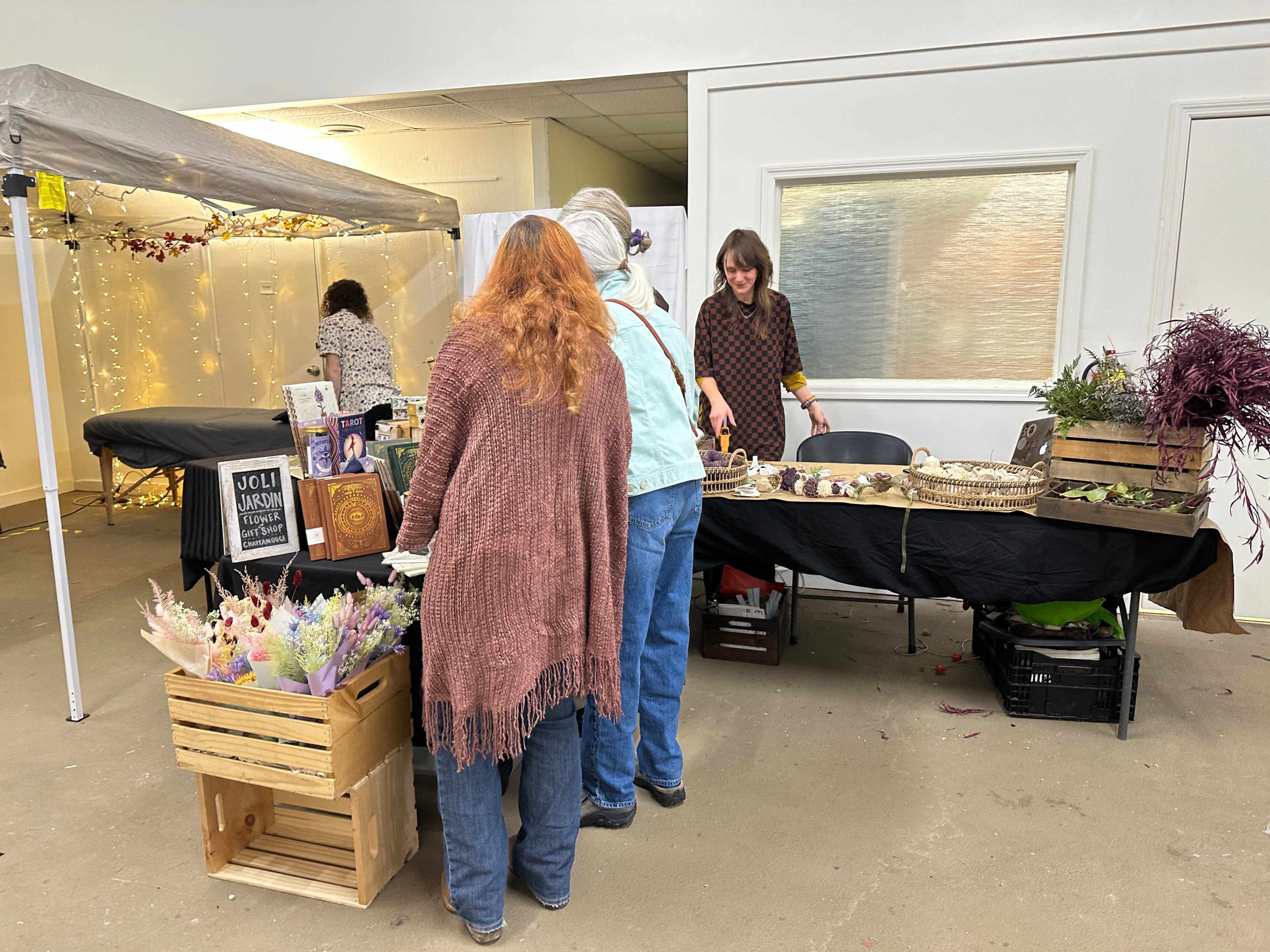 Two people browse a craft vendor booth adorned with dried flowers and handmade items at a market.