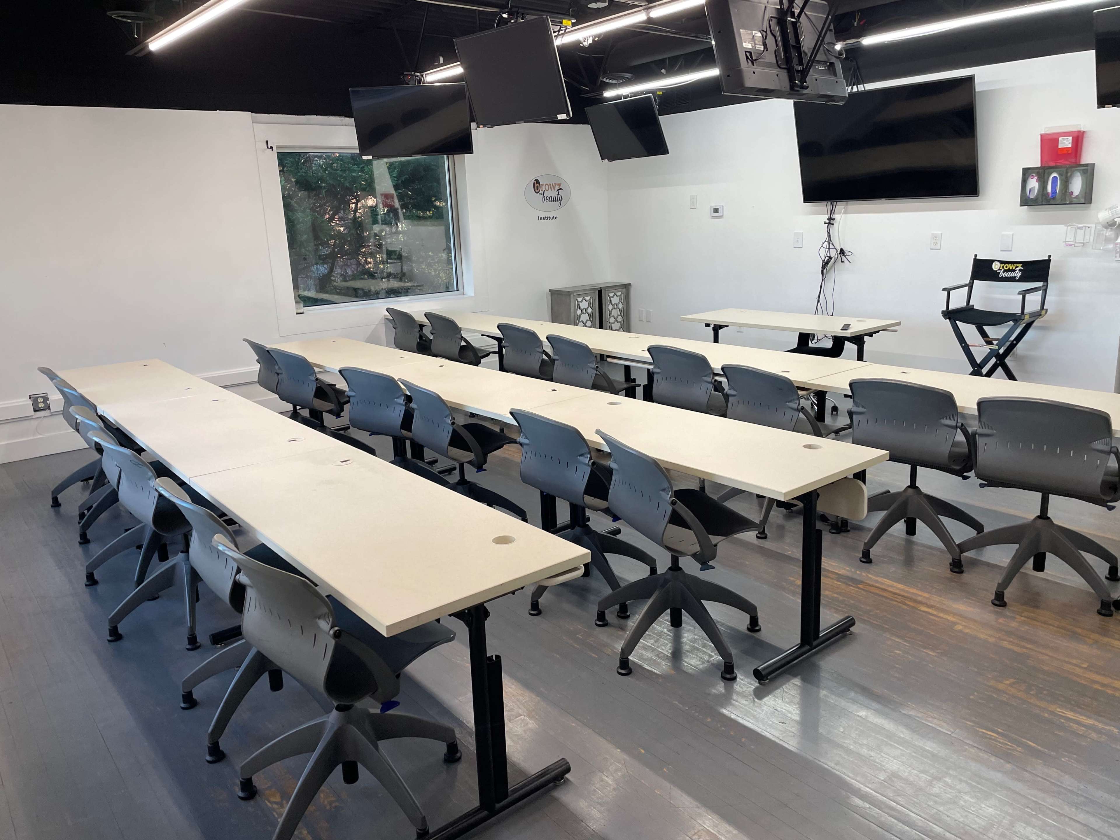 A spacious classroom setting with multiple rows of long tables and rolling chairs, equipped with monitors on the walls.