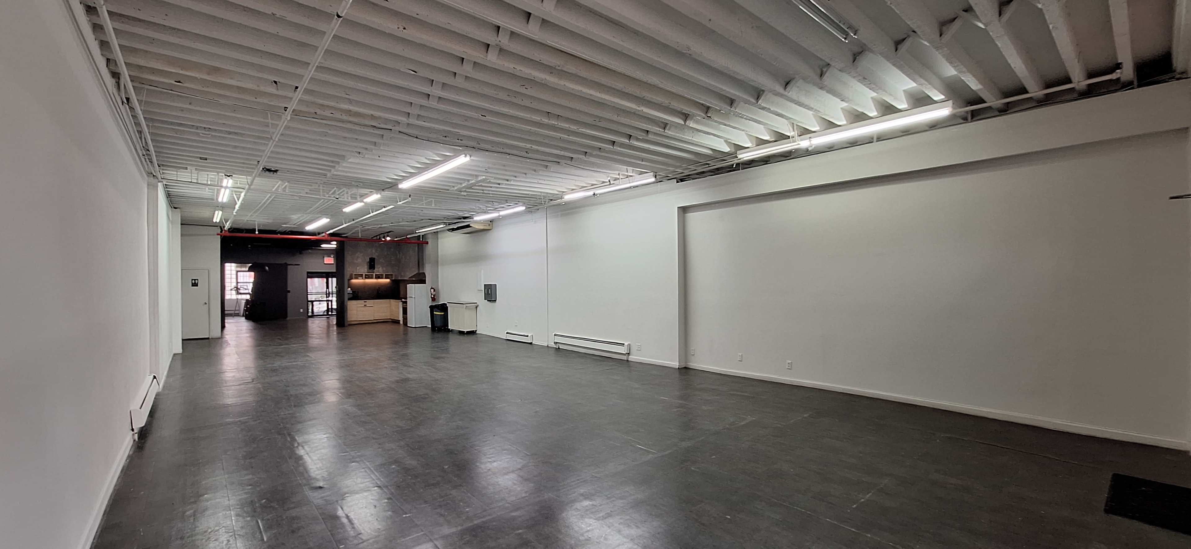 The image shows a spacious, empty room with a smooth floor, white walls, and fluorescent lighting, leading into another room visible in the background.