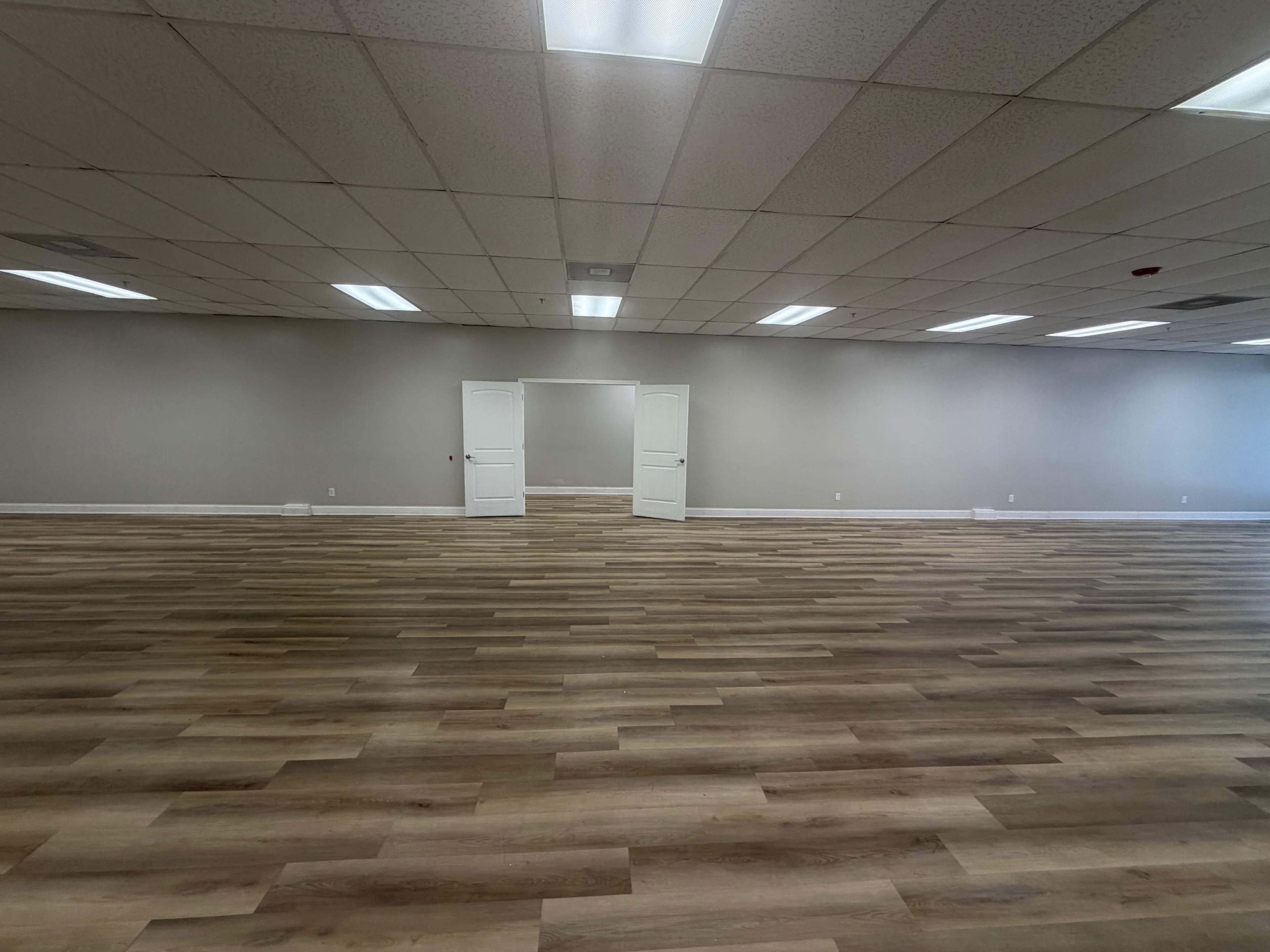 The image shows a spacious, empty room with hardwood-like flooring and double doors leading to another area in the back.