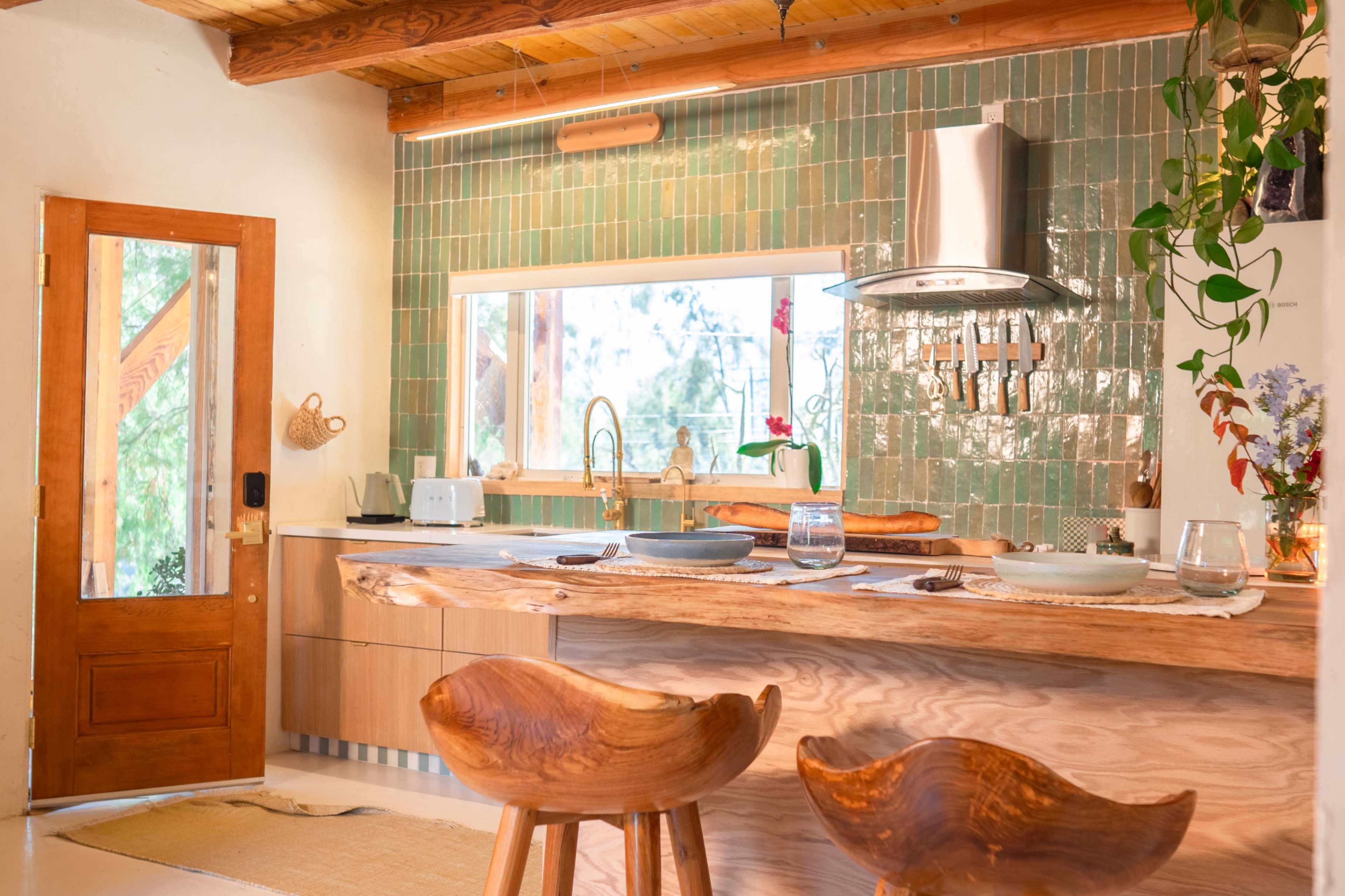 The image shows a modern kitchen featuring wooden cabinetry, green tiled walls, and a large window allowing natural light to enter.
