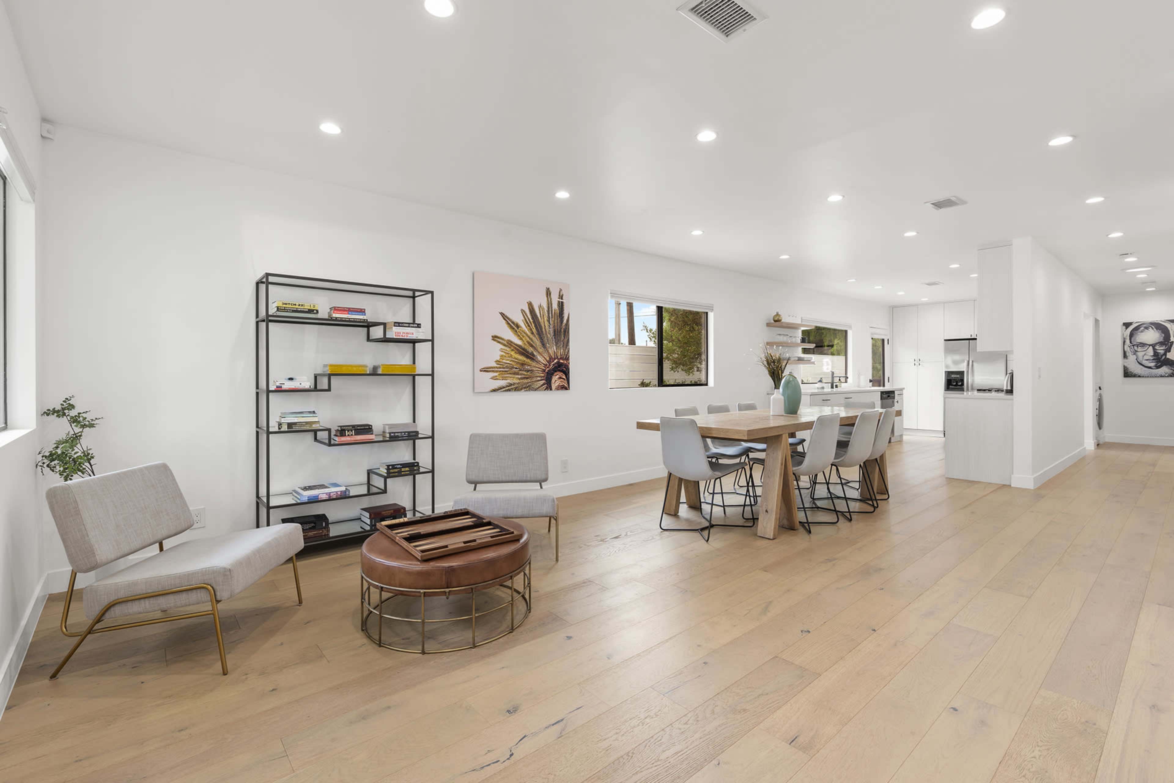 A spacious, modern living area features a dining table with six chairs, a bookshelf, and large windows allowing natural light.
