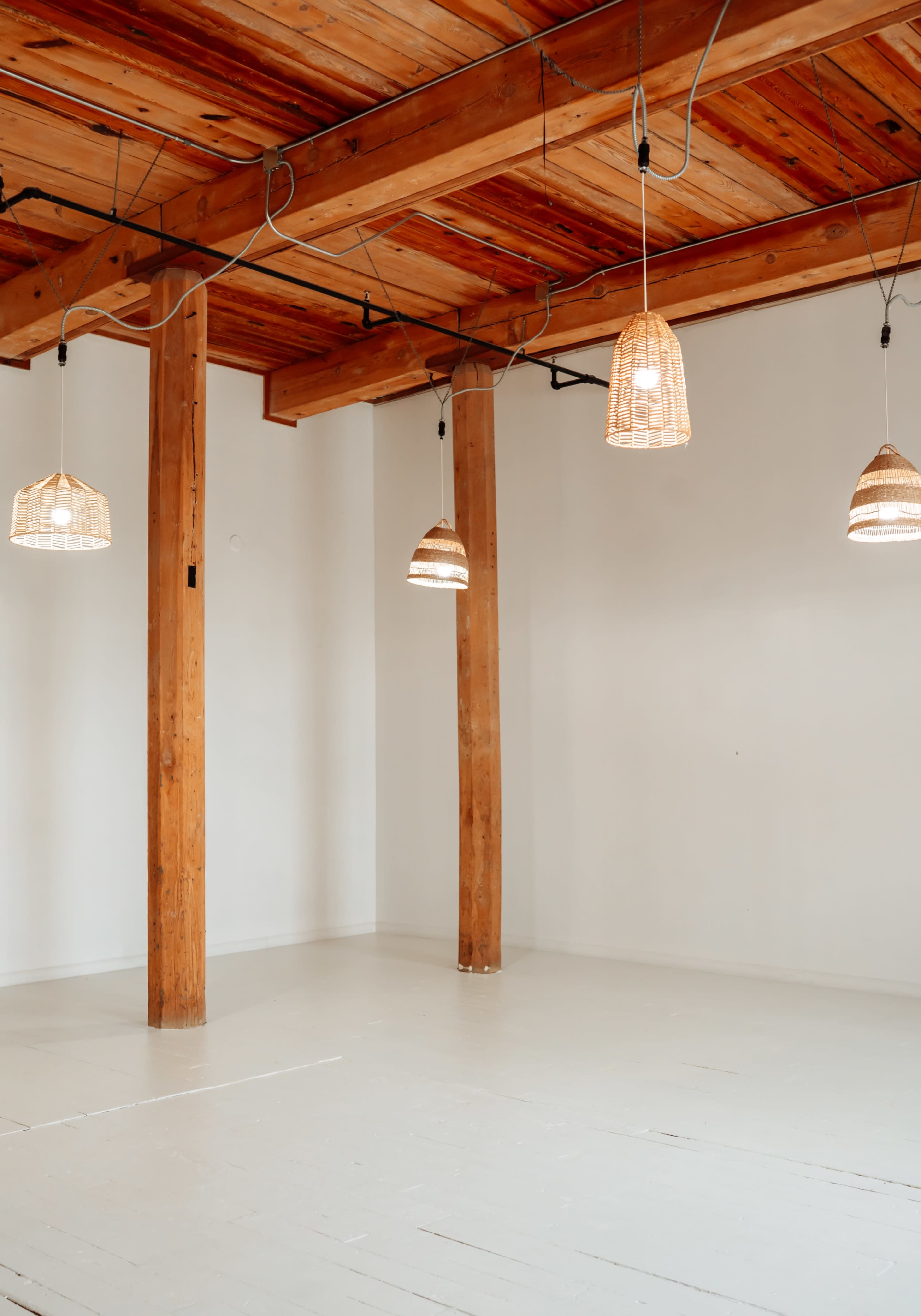 The image shows a spacious room with wooden beams and pendant lights hanging from the ceiling, along with a white-painted floor.