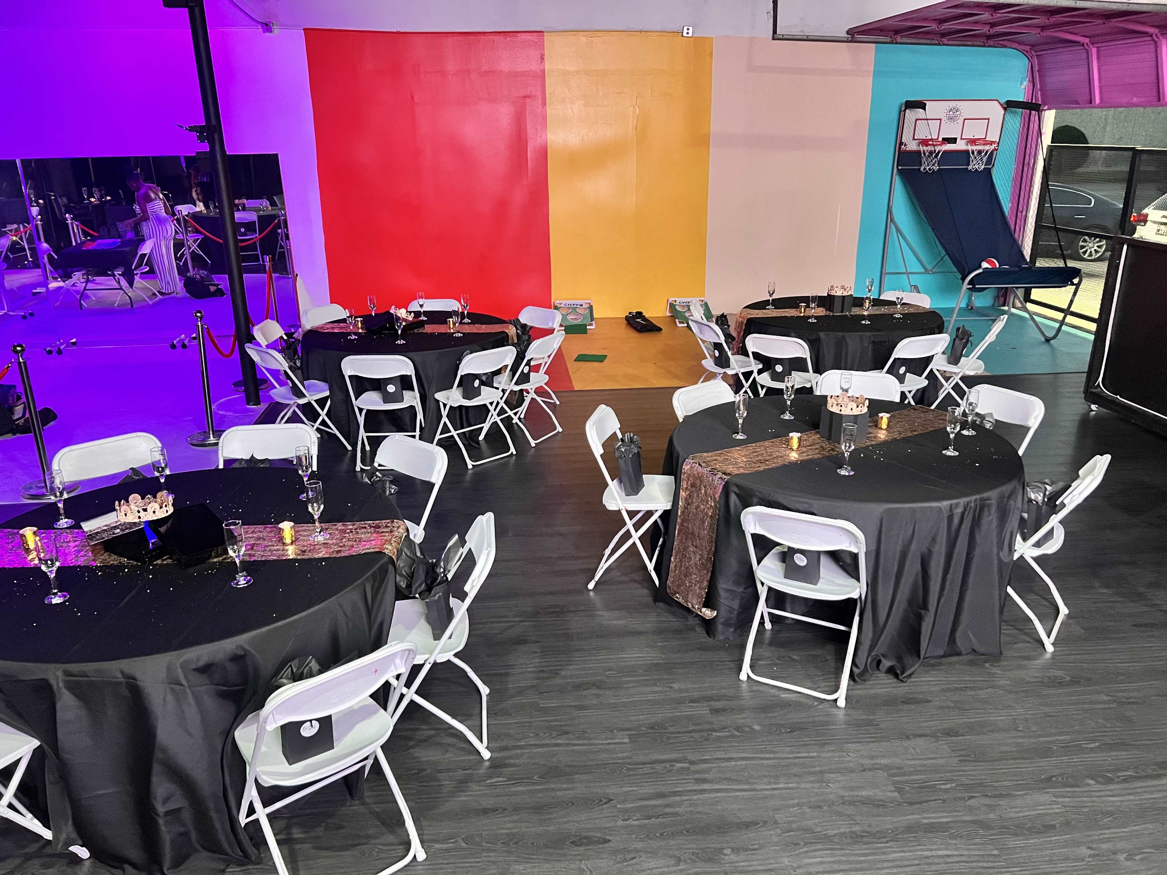 A party setup with several round tables covered in black tablecloths, accompanied by white chairs, in a brightly colored indoor space.