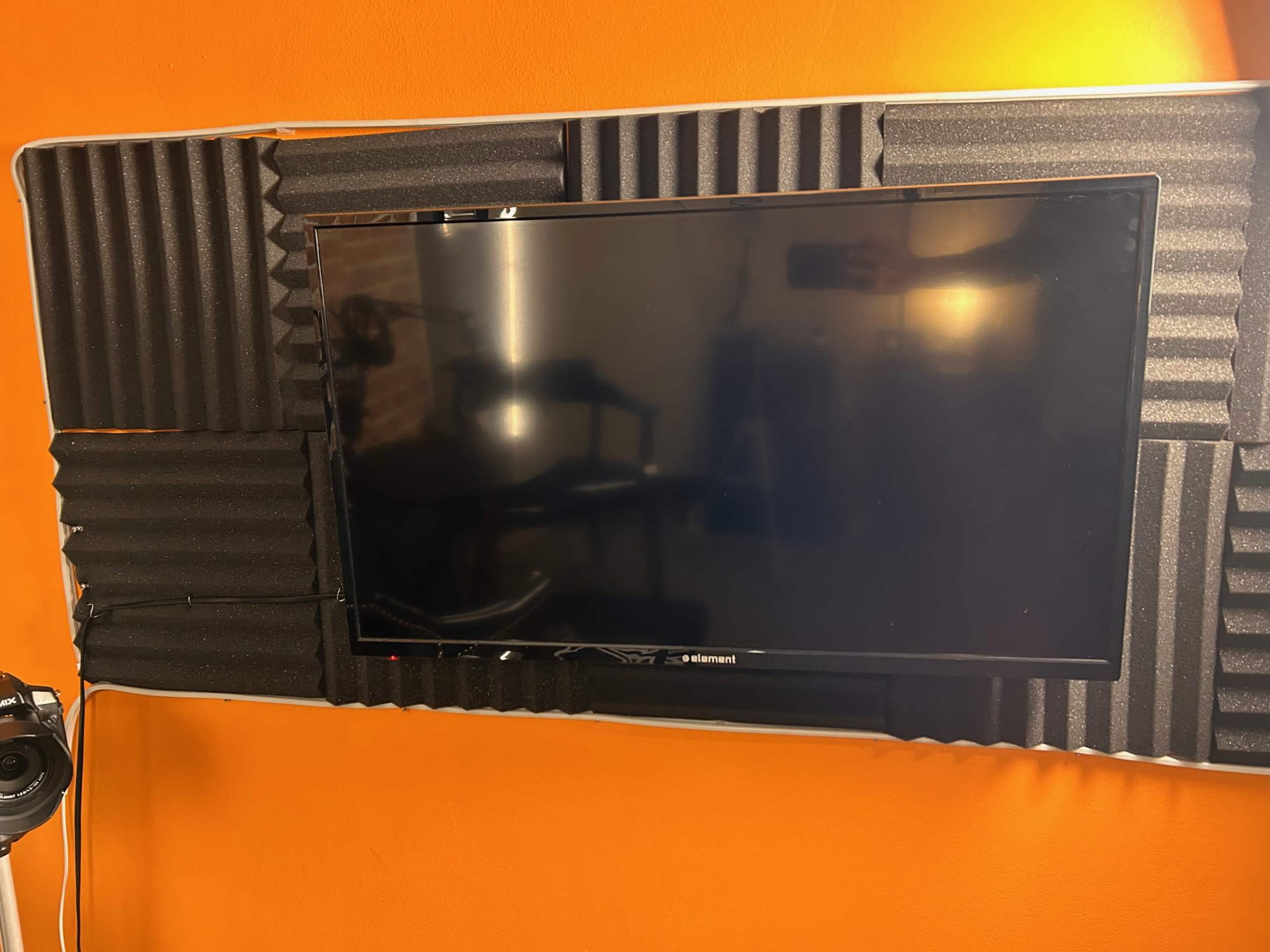 A black television is mounted on a wall covered with gray soundproofing foam, against an orange backdrop.
