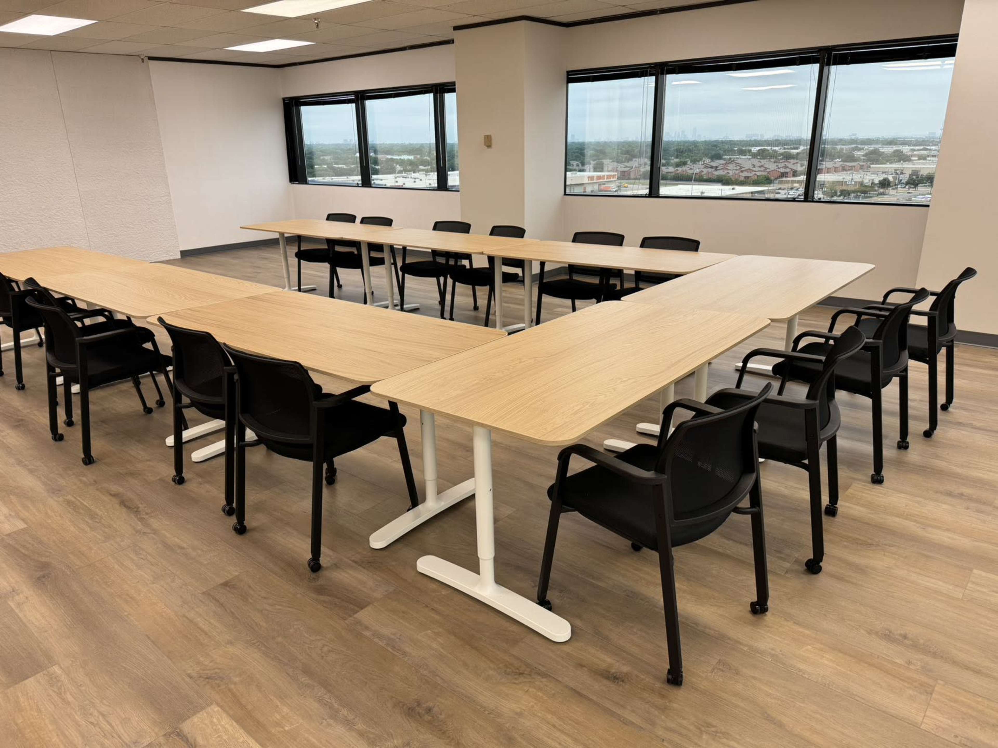 A large meeting room features a U-shaped arrangement of wooden tables surrounded by black chairs and large windows.