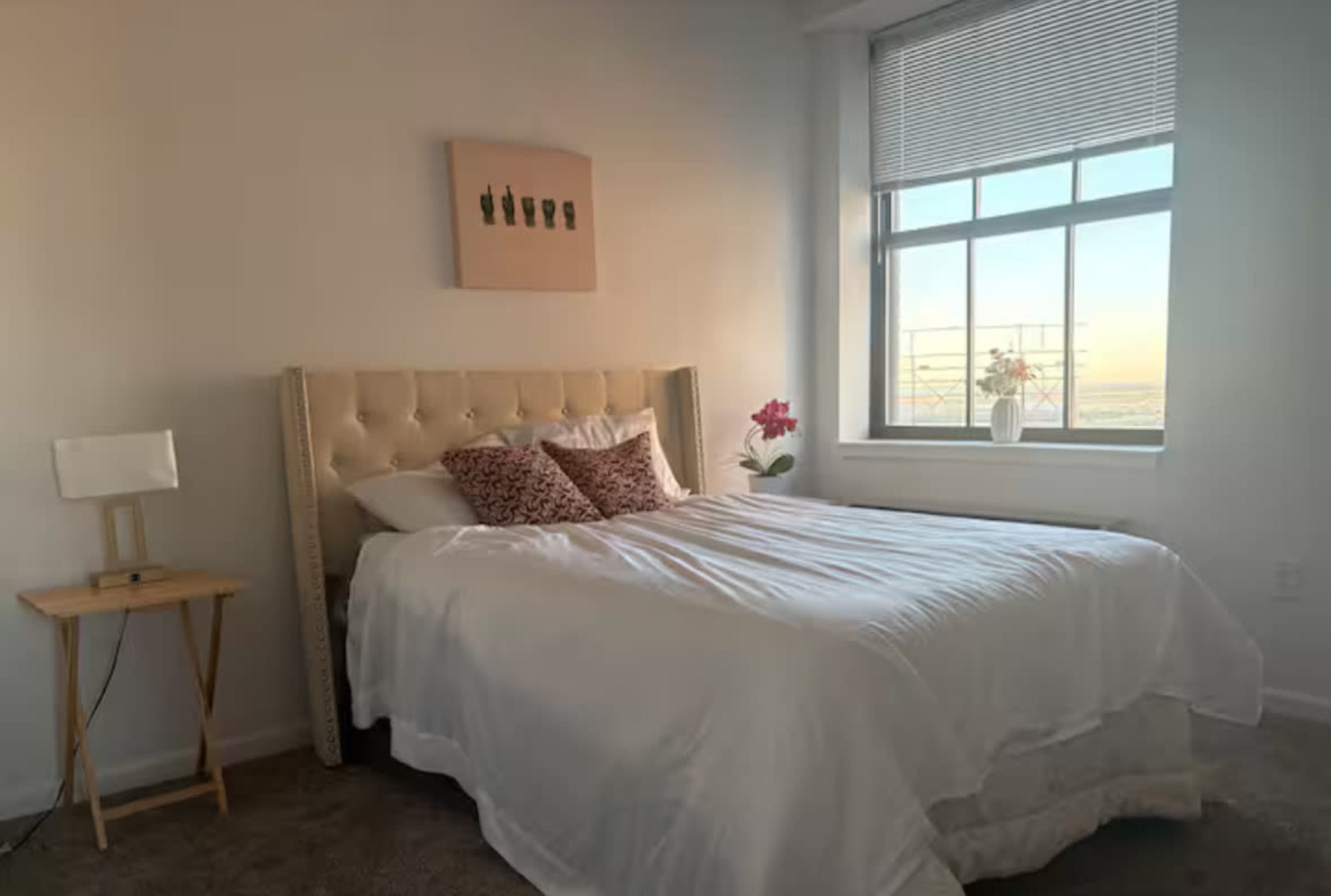 The image shows a bright bedroom featuring a large bed with a light-colored comforter, decorative pillows, and a window with blinds, alongside a small bedside table.