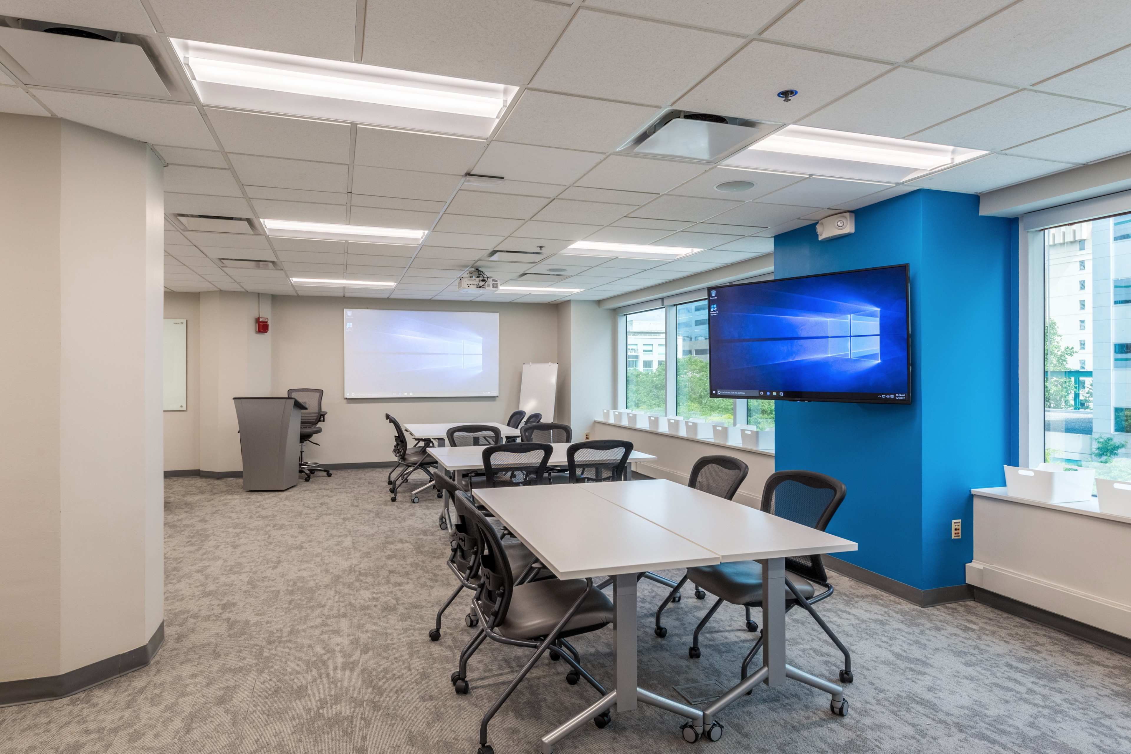 A modern conference room features a large screen, multiple tables, and ergonomic chairs, with natural light coming through the windows.