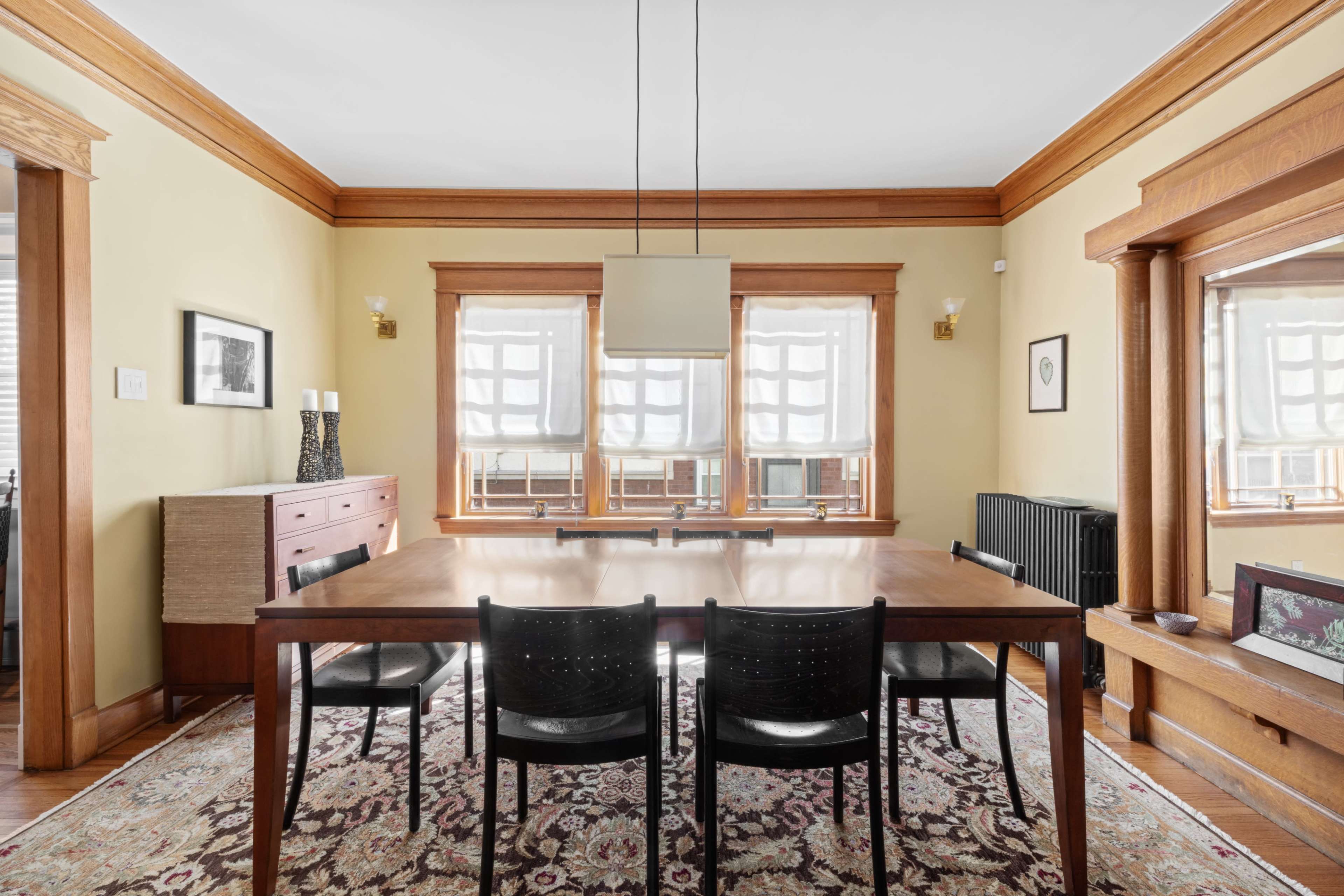 A dining room features a large wooden table surrounded by black chairs, with windows and a sideboard visible in a well-lit space.