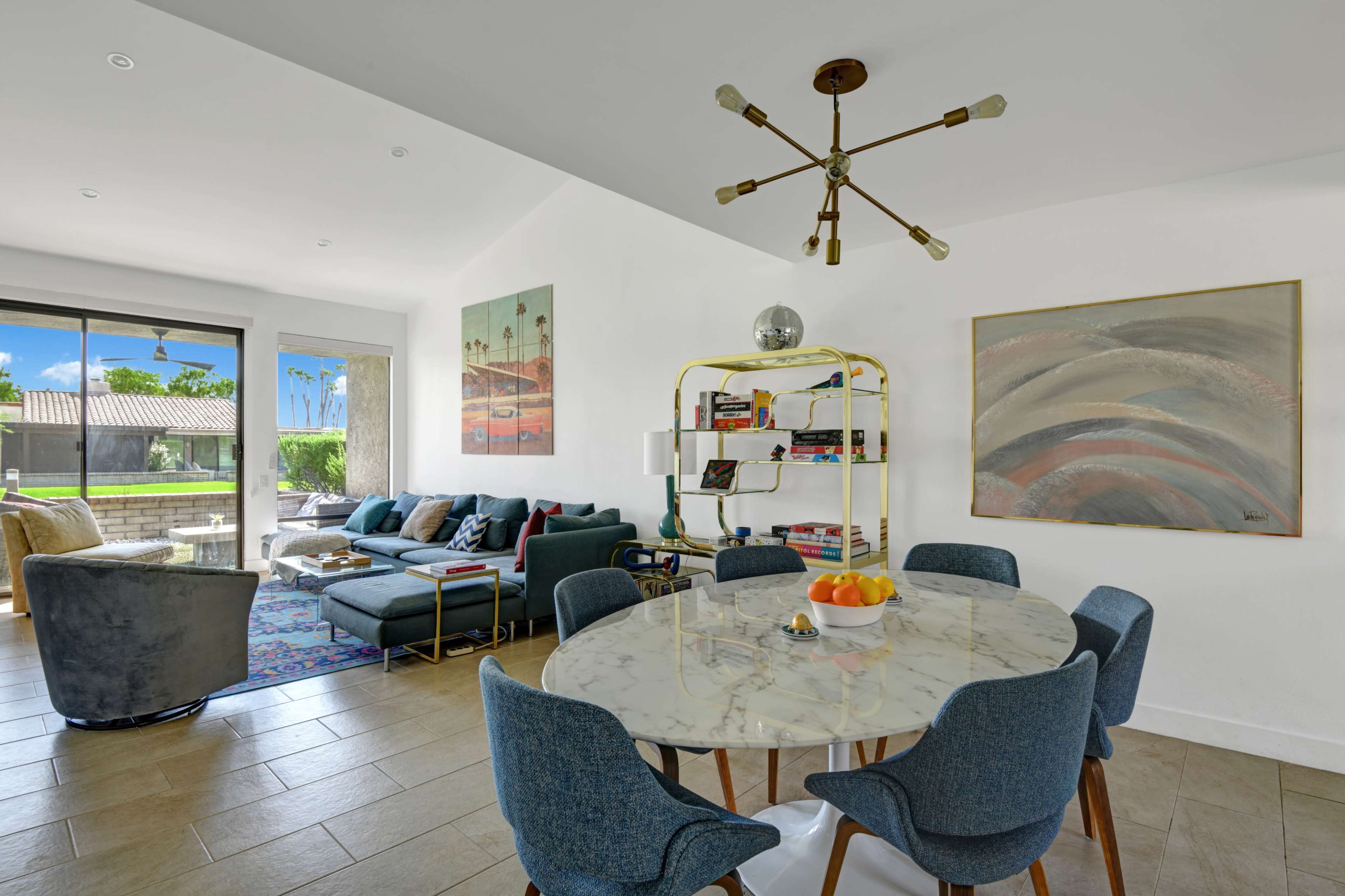 A modern dining area features a round marble table surrounded by blue chairs, with a living space and large windows visible in the background.