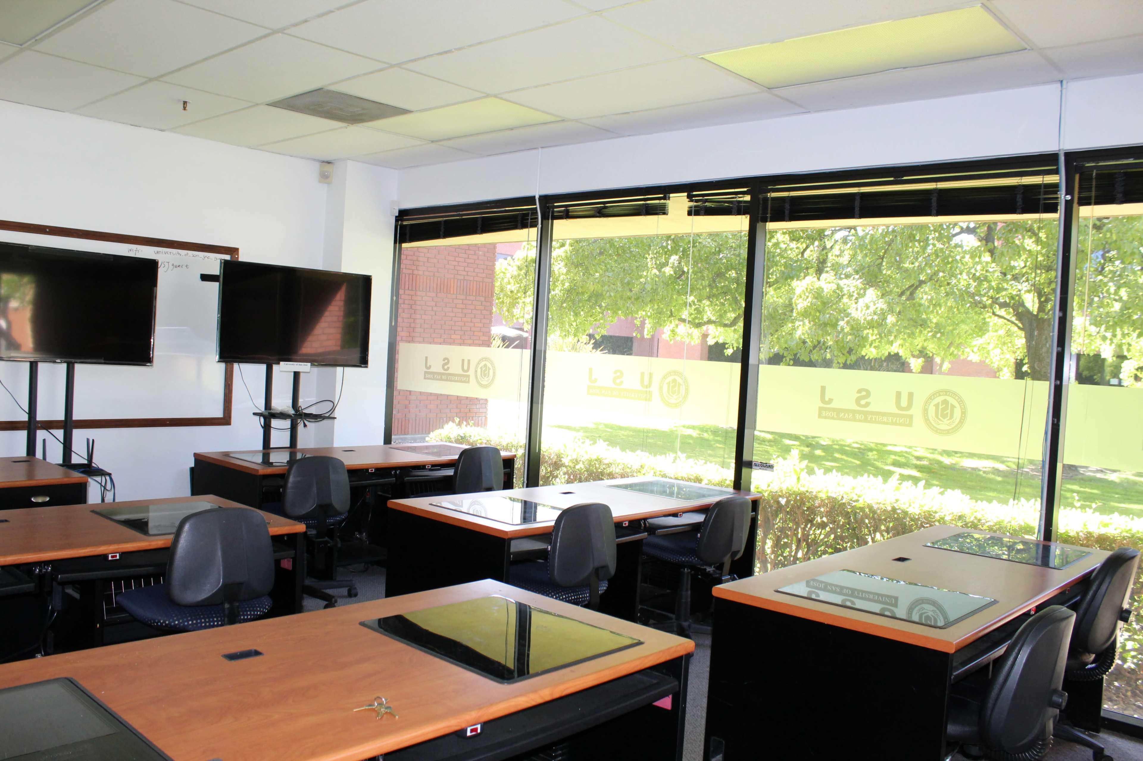 A classroom is equipped with desks and chairs, featuring large windows that overlook greenery outside.