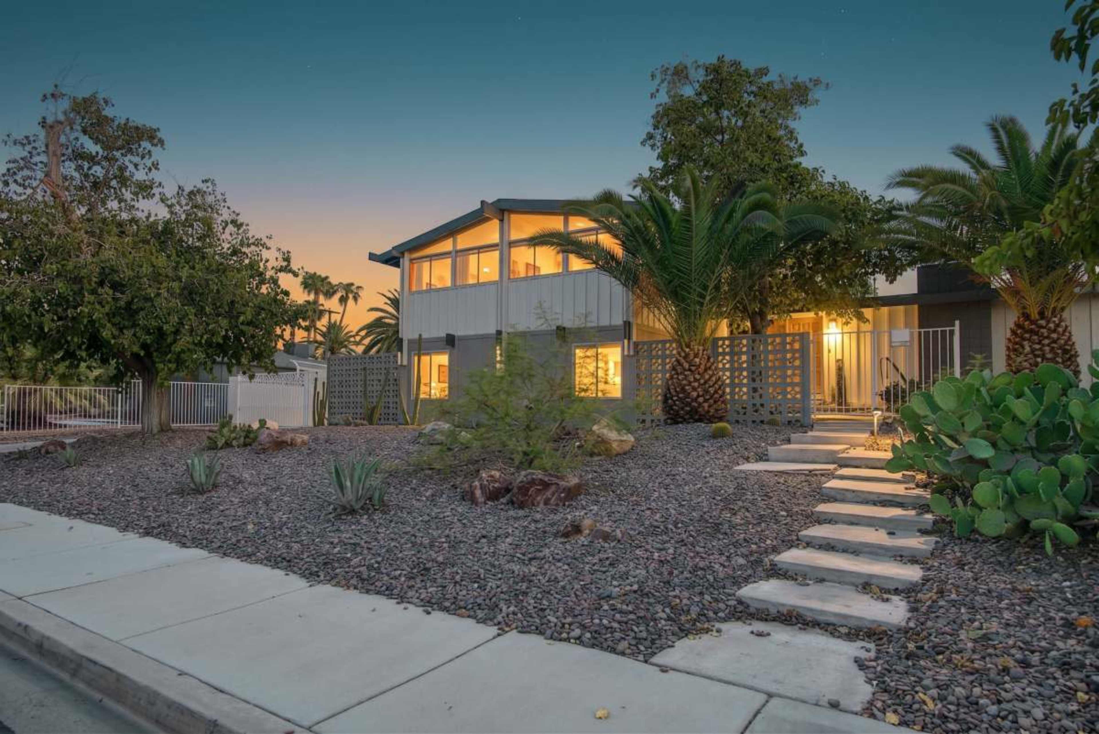 The image shows a modern house with large windows, surrounded by a landscaped yard featuring rocks, plants, and palm trees.