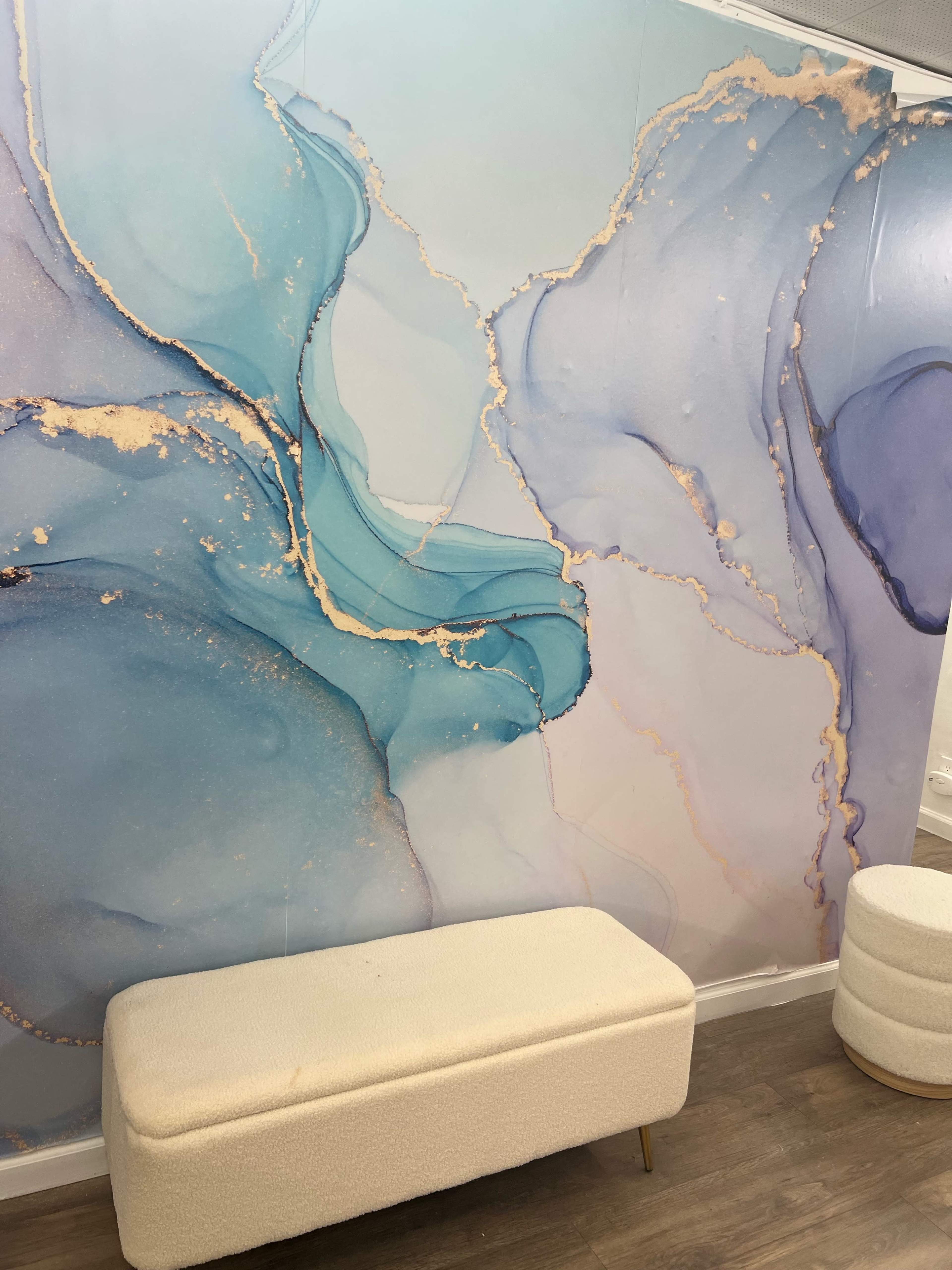 A textured wall with a blue and purple abstract design and a light-colored, upholstered bench in front of it.