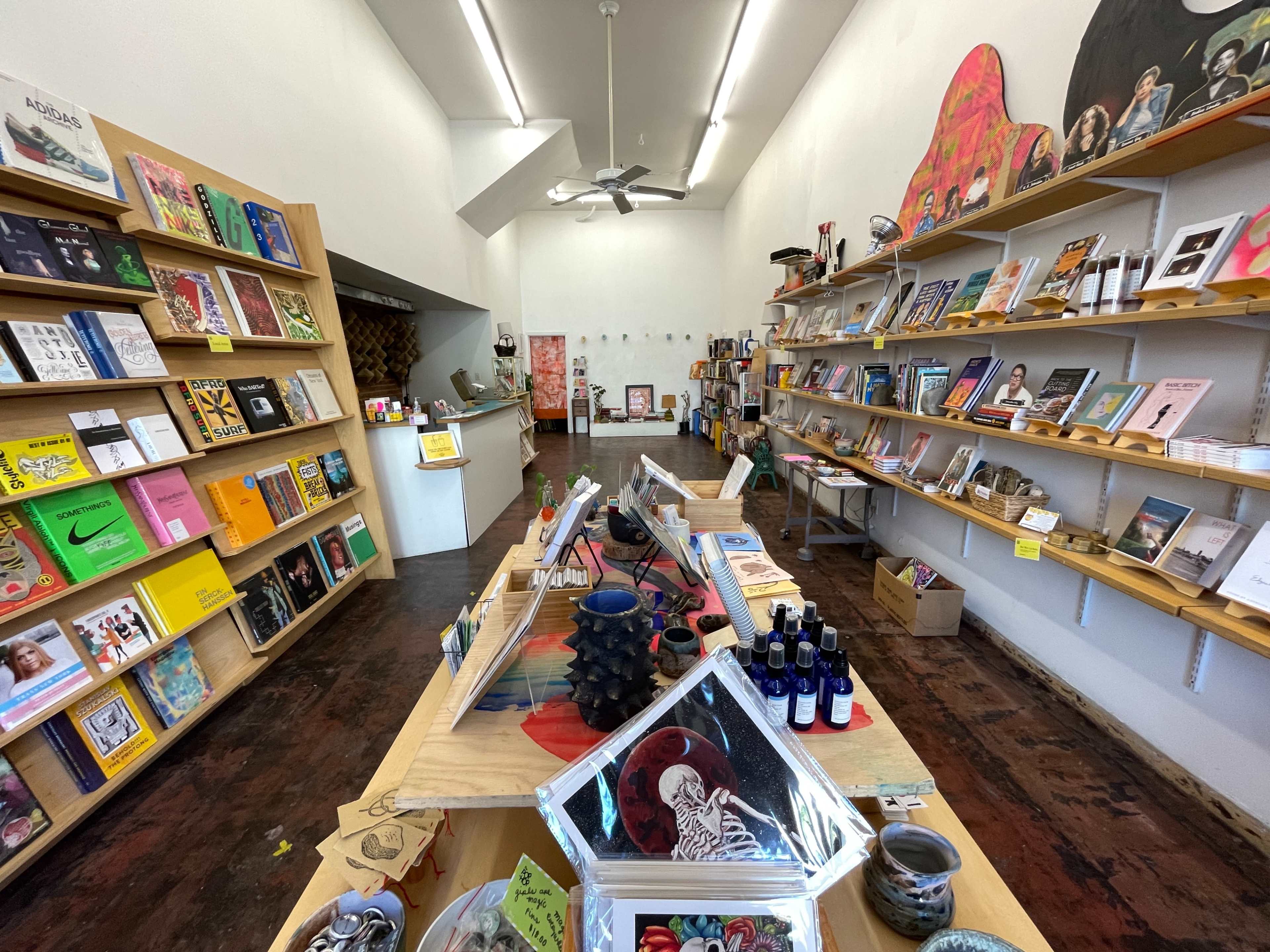 The image shows the interior of a bookstore with wooden shelves displaying various books and art products, and a checkout counter in the foreground.