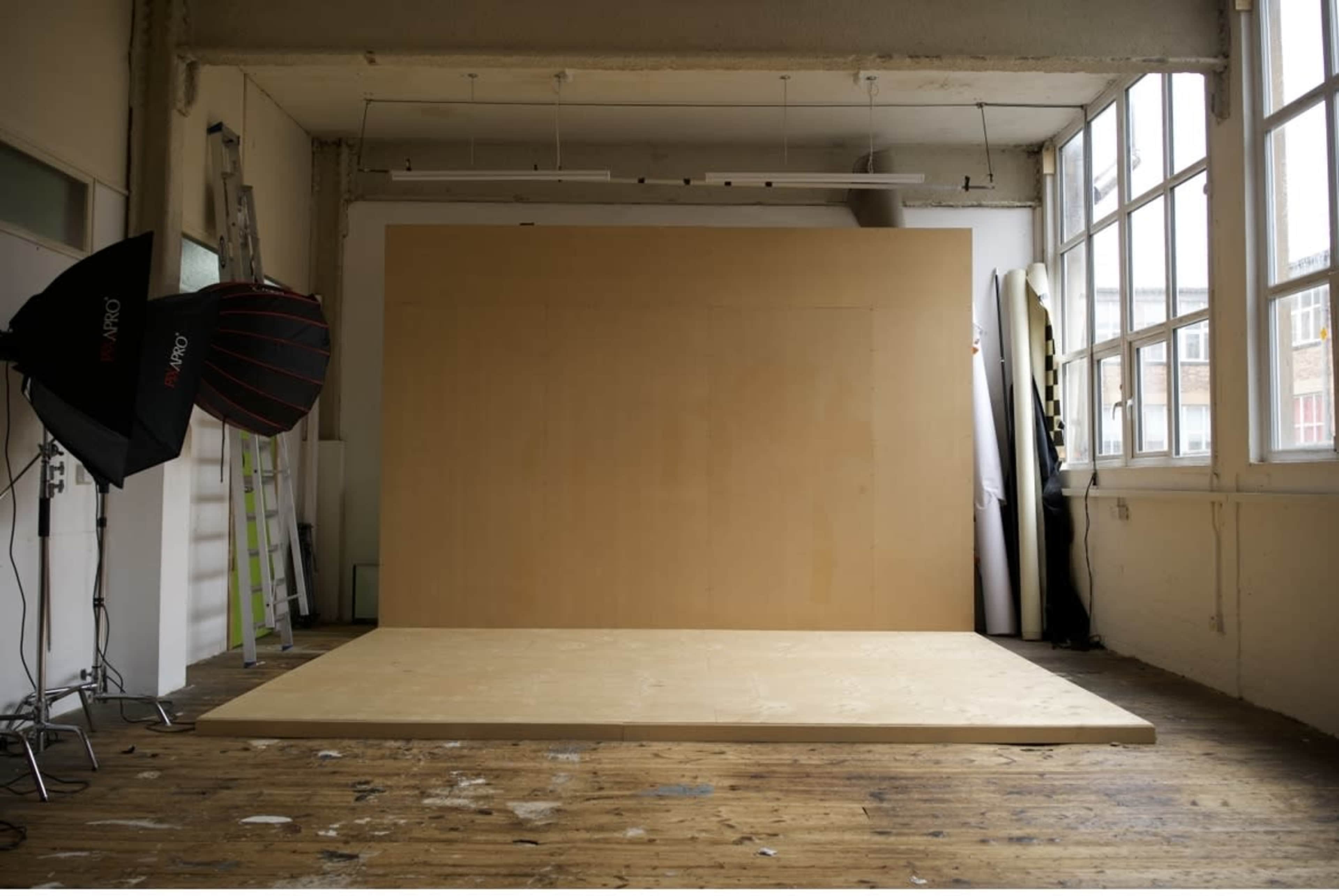 A spacious photo studio with a wooden platform and a large blank wall, illuminated by natural light from the windows.