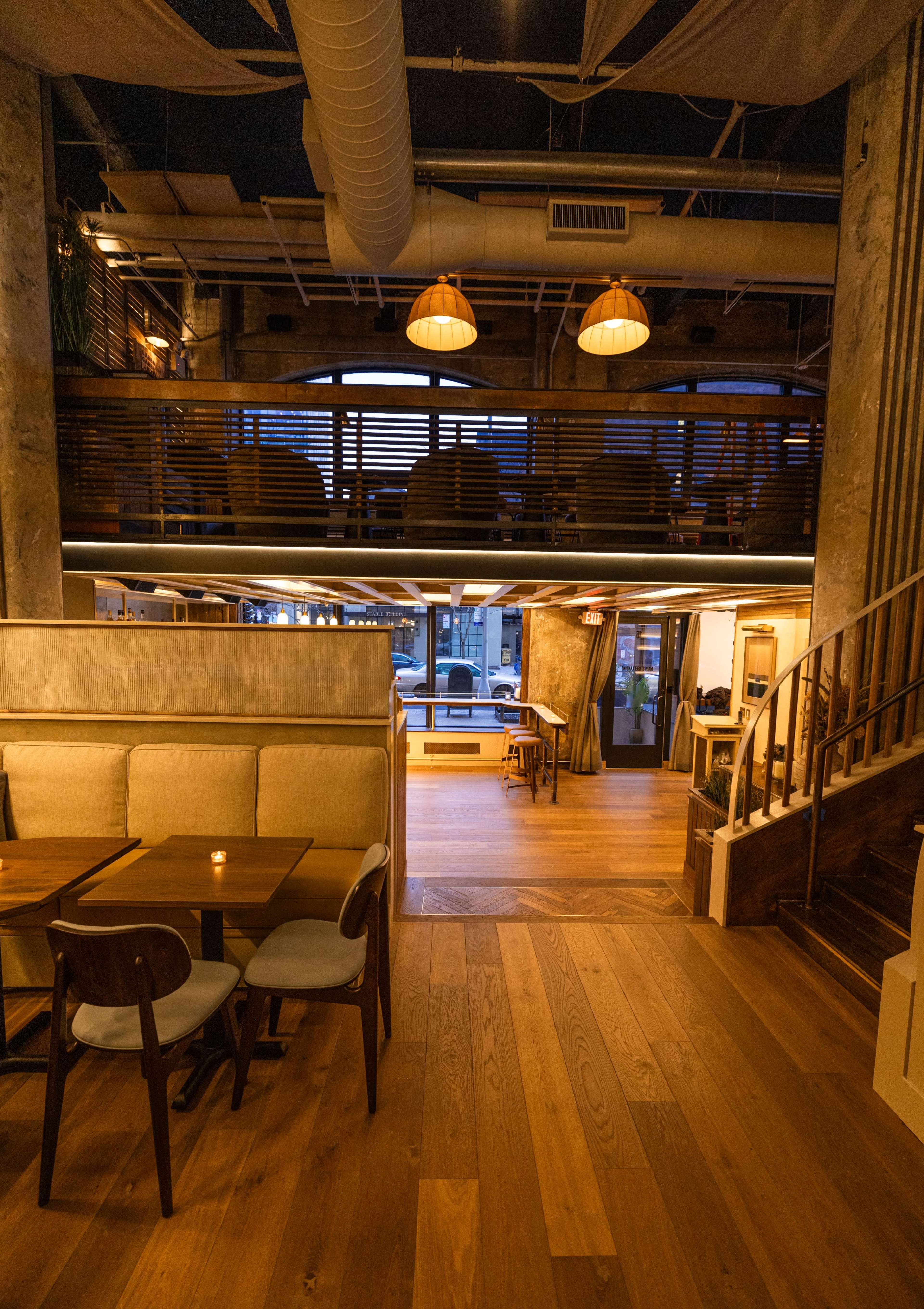 The interior of a restaurant, featuring wooden flooring, seating areas with tables, and a two-level layout illuminated by hanging lights.