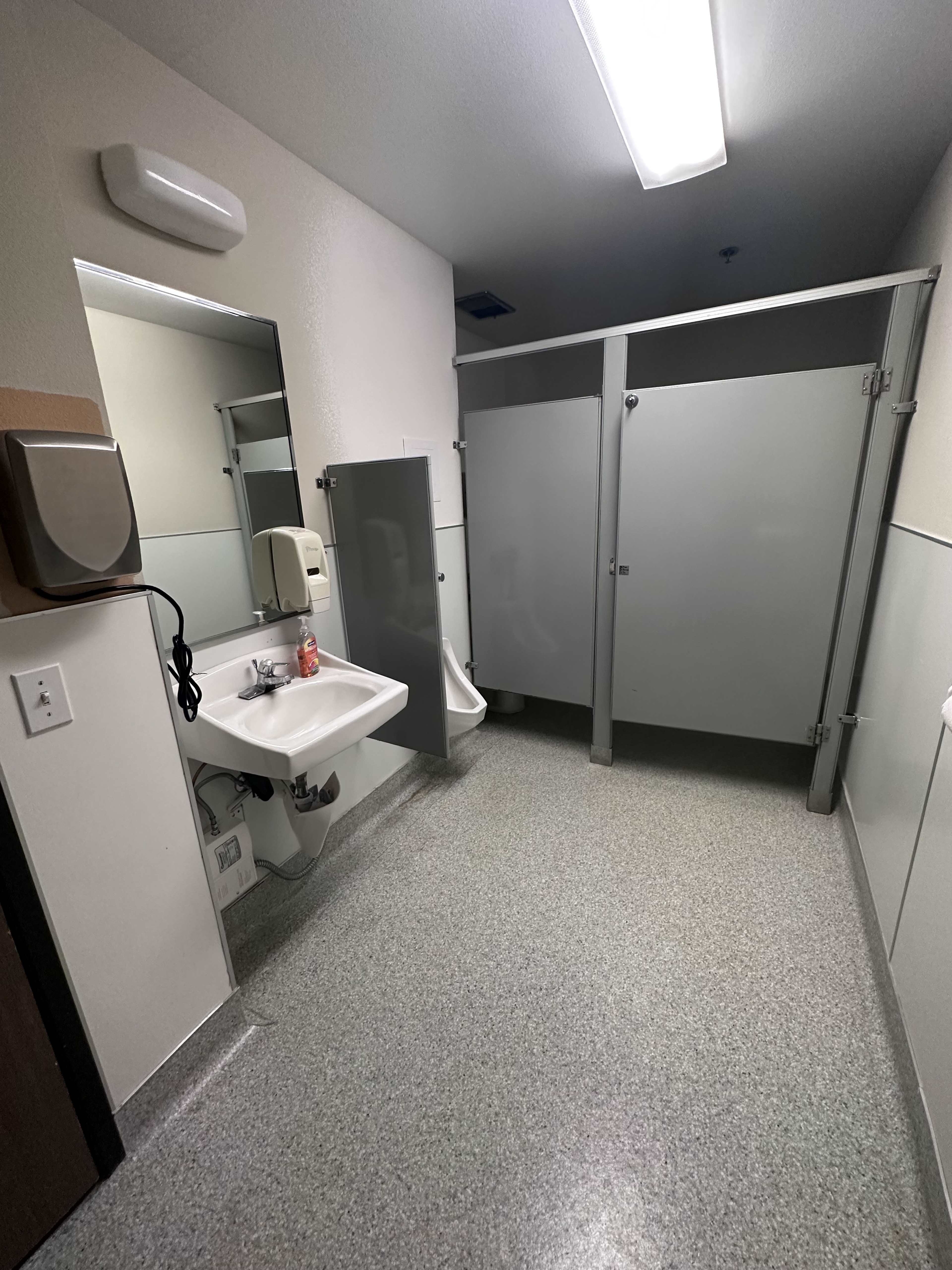 The image shows a restroom with a sink, a hand dryer, a mirror, and three partitioned stalls.