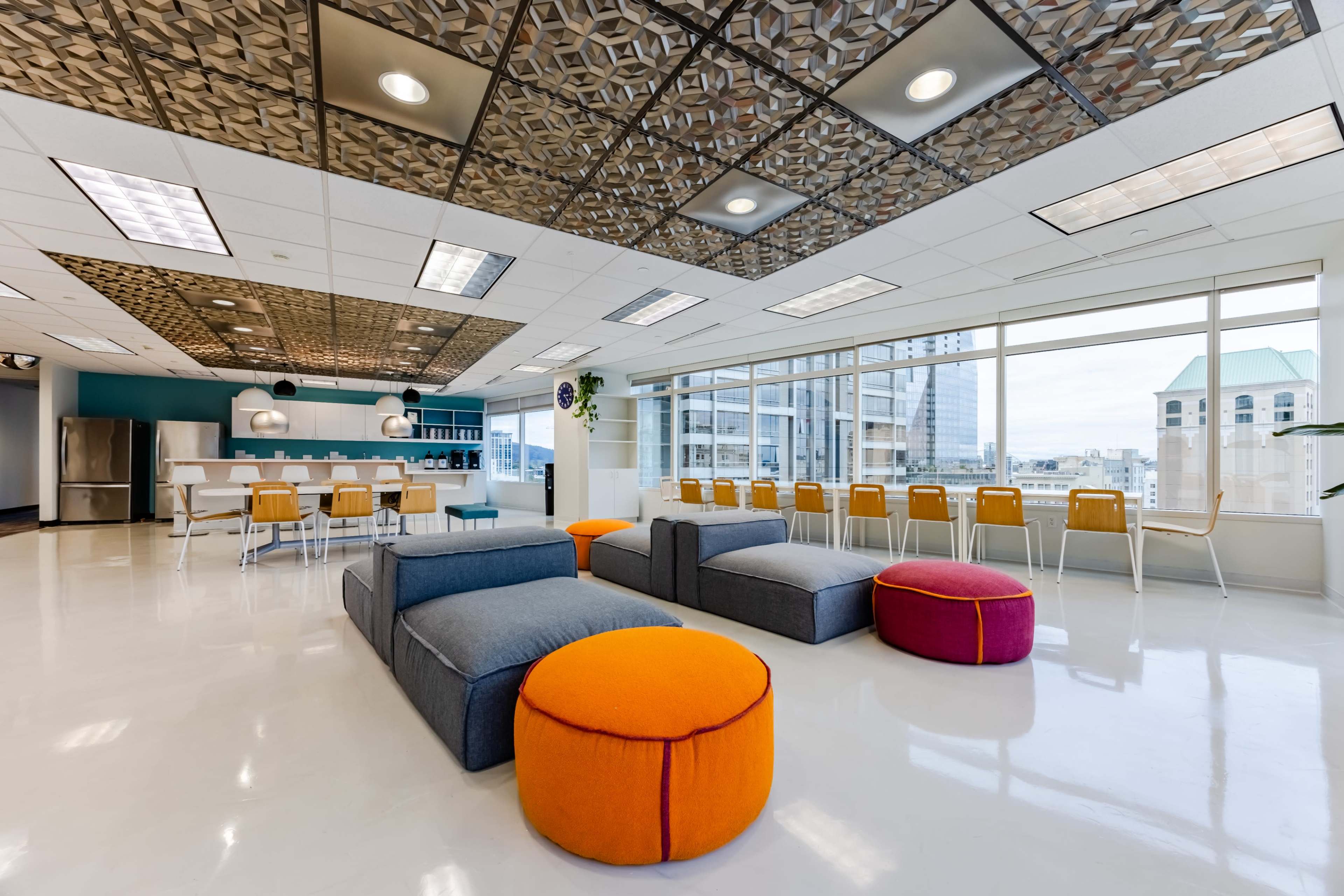 The image shows a modern office lounge with spacious seating areas, a kitchen counter, and large windows allowing natural light.