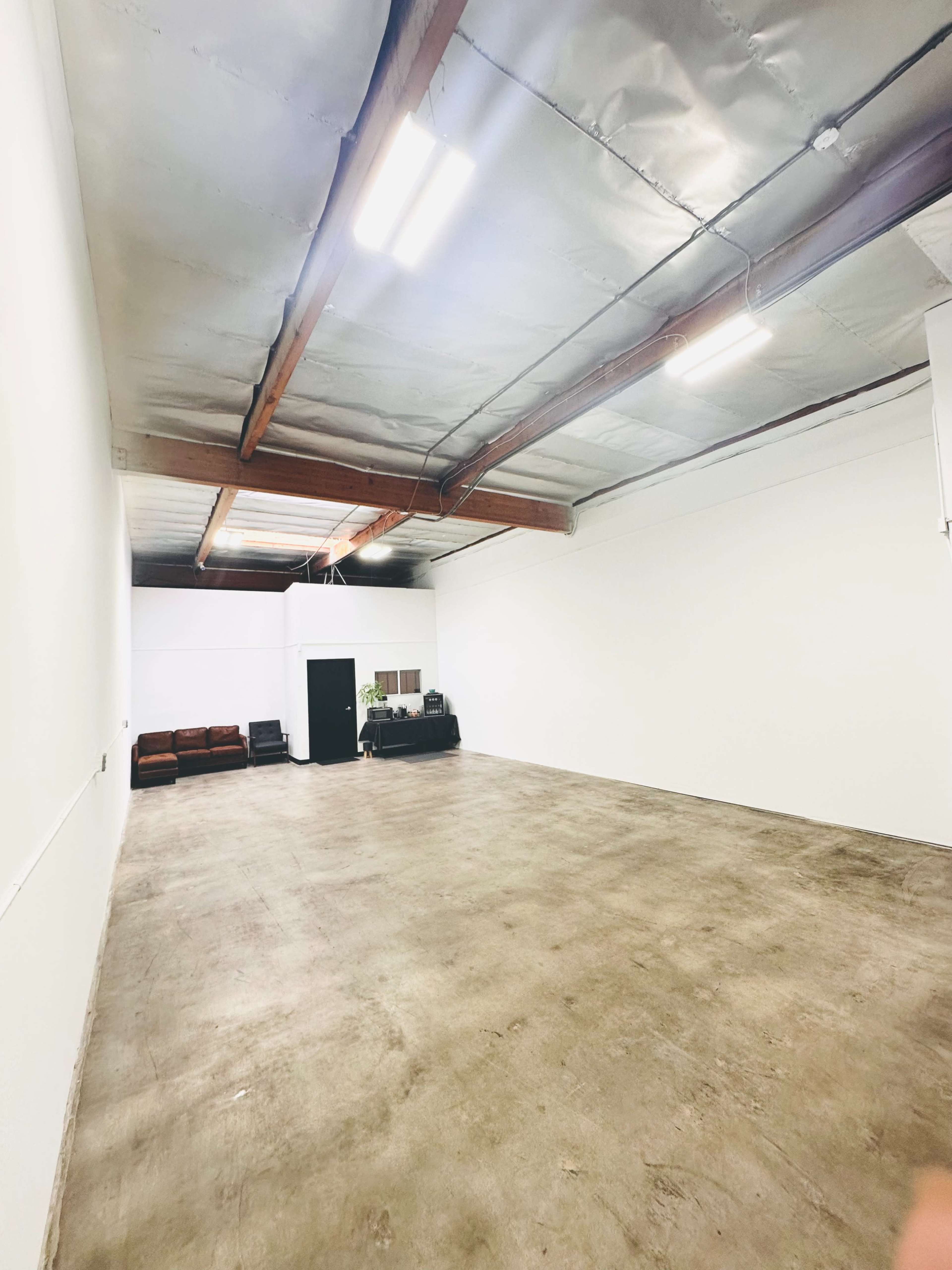 An empty room with concrete flooring, exposed wooden beams, and minimal furnishings, including a sofa and a black door at the far end.