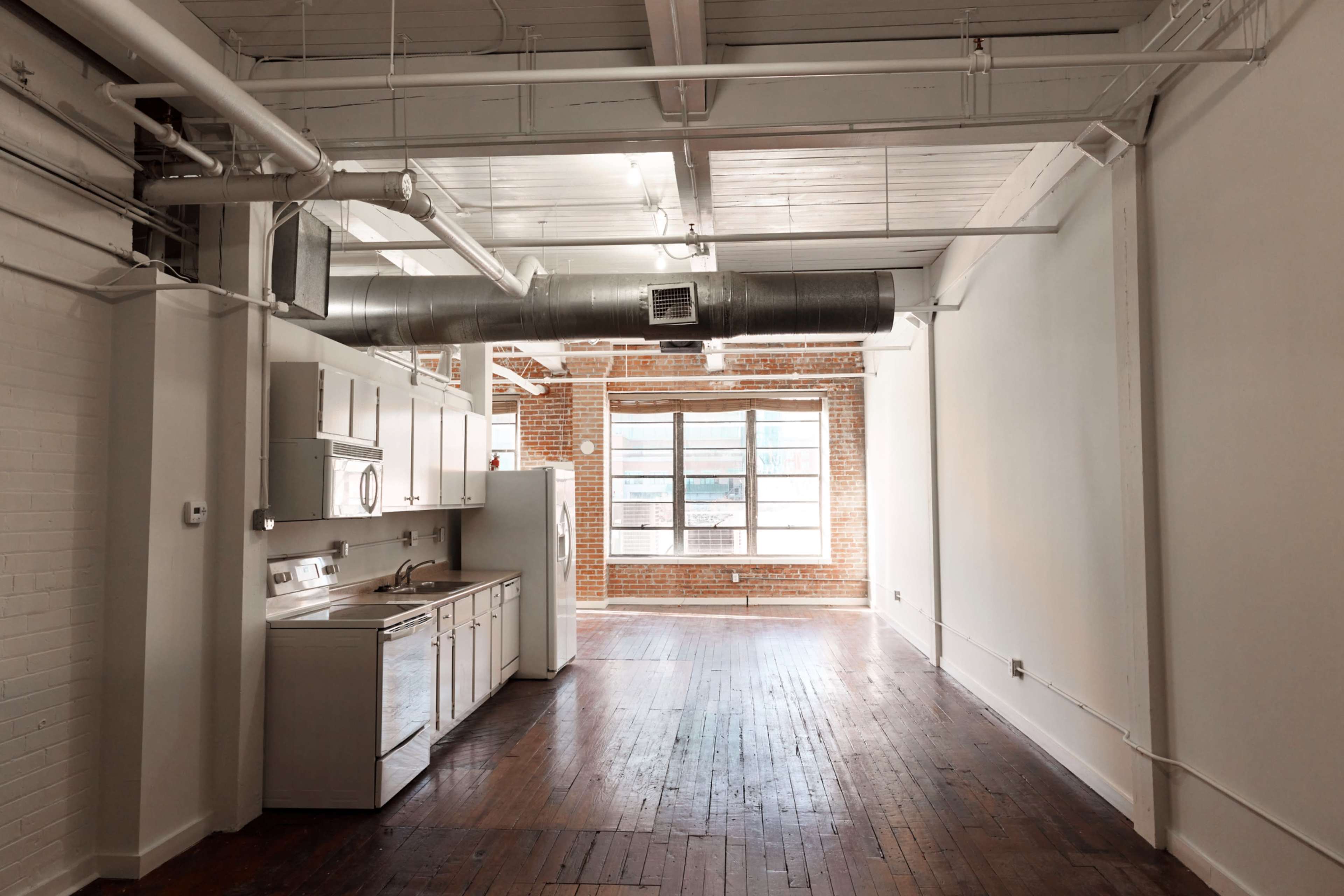 An open loft space with hardwood floors, exposed brick walls, and a kitchen area featuring white cabinetry and appliances.