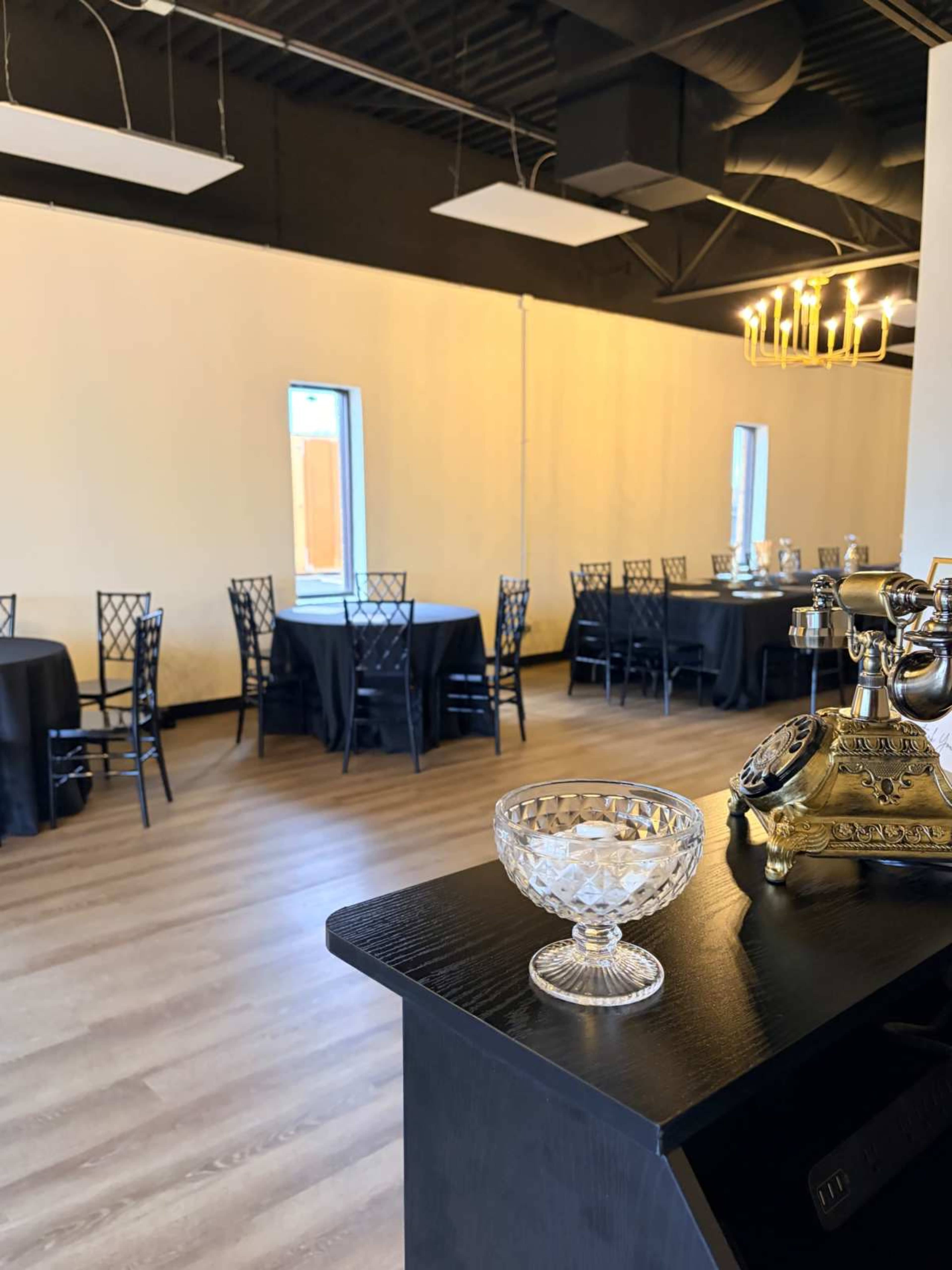 The image shows a spacious, partially furnished event room with black tables and chairs arranged for a gathering, featuring a decorative glass bowl and a vintage telephone on a counter in the foreground.