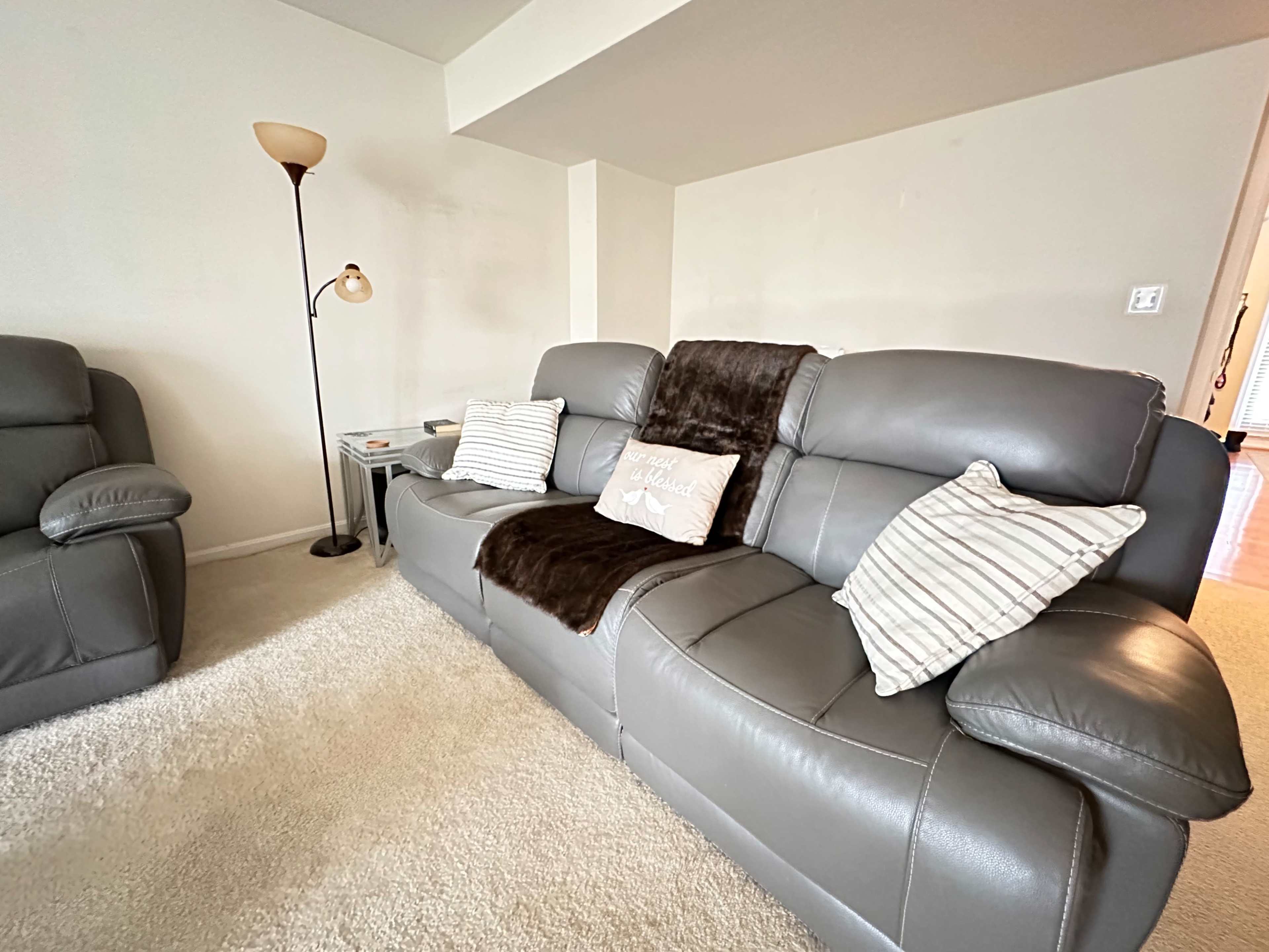 A gray leather sofa with decorative pillows and a throw blanket is positioned in a well-lit living room beside a floor lamp and side table.
