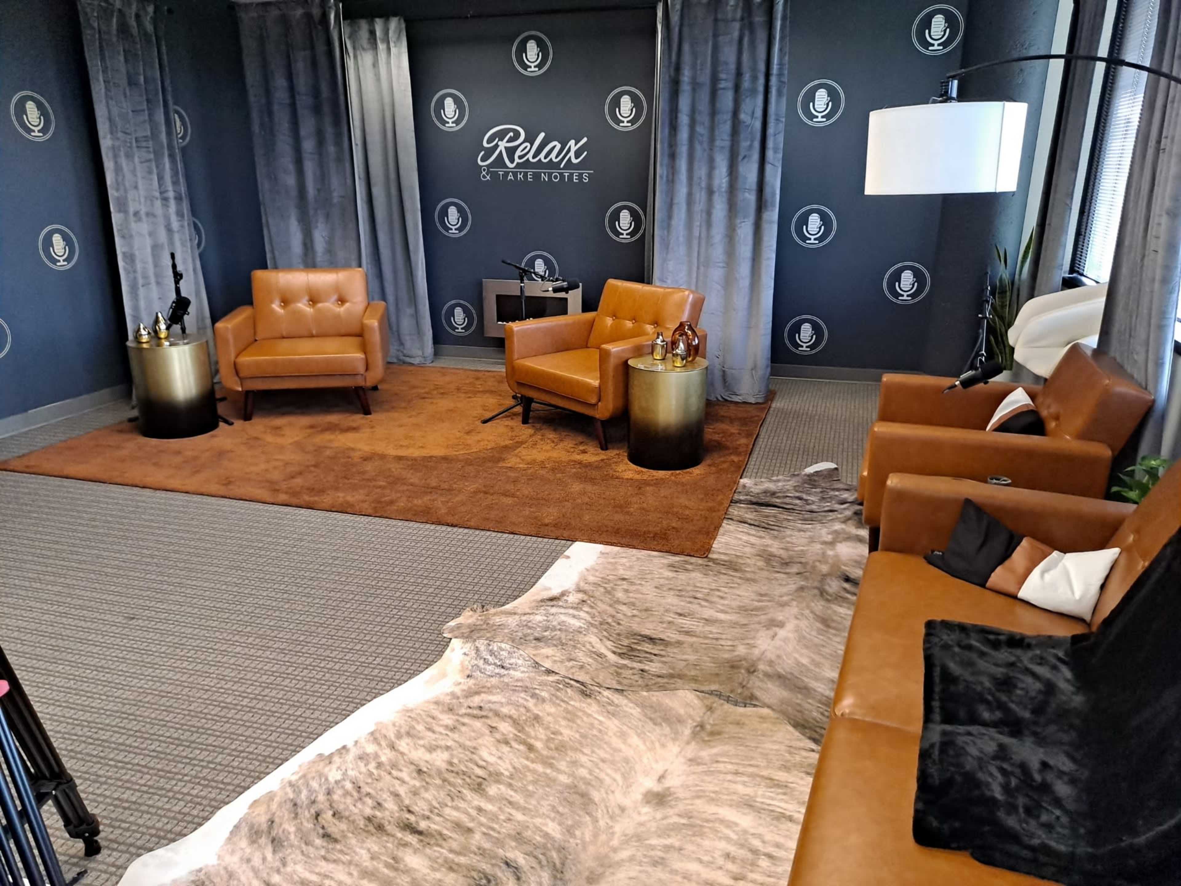 A modern, styled interview set featuring two brown leather chairs, a round side table, a microphone stand, and a large area rug on a carpeted floor.