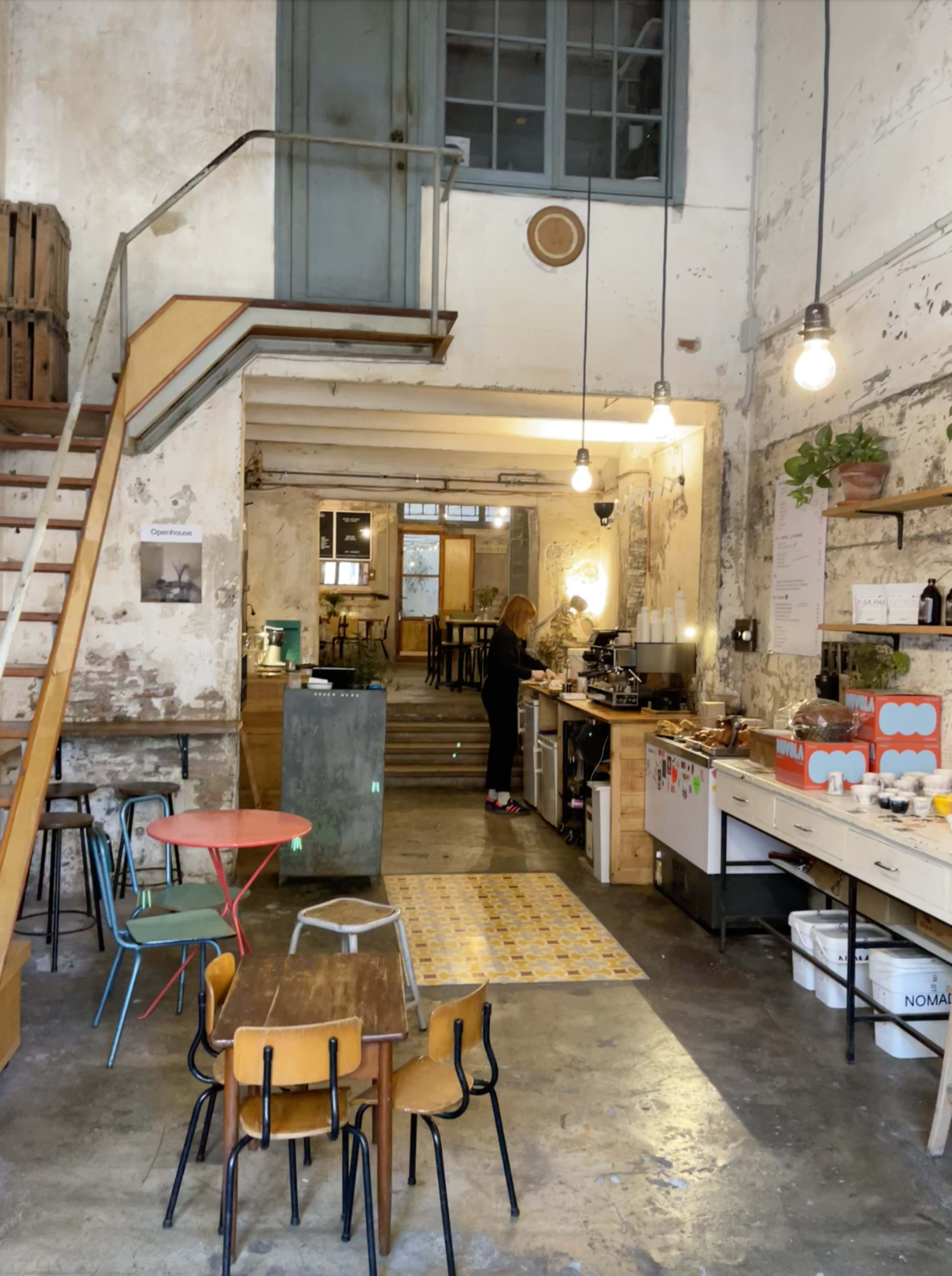 A spacious, rustic café with a staircase on the left, a woman preparing coffee at the counter, and various seating arrangements scattered throughout the room.