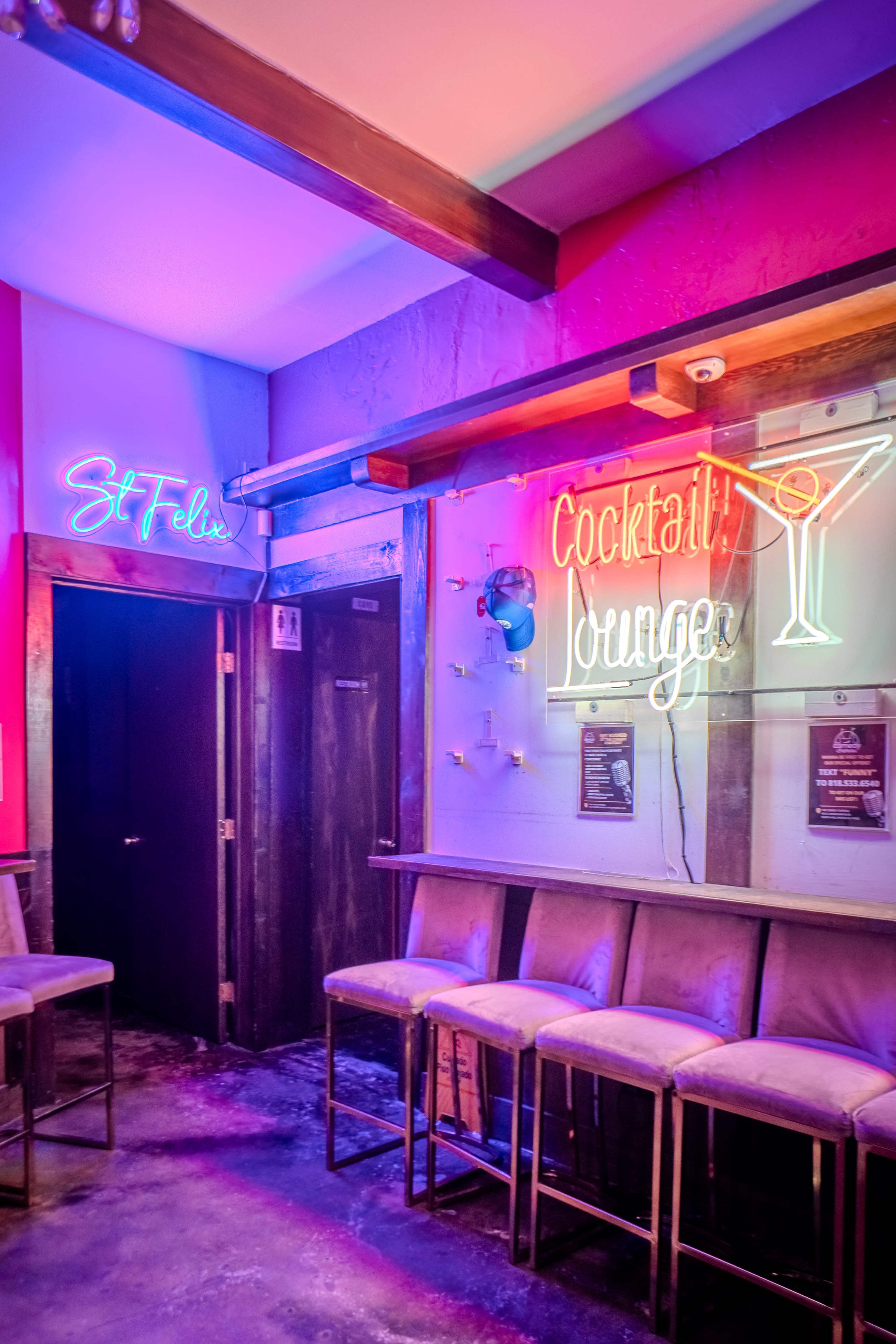 The image shows a dimly lit bar area with neon signs reading "St Felix" and "Cocktail Lounge" on opposite walls, along with empty high stools lined against a counter.