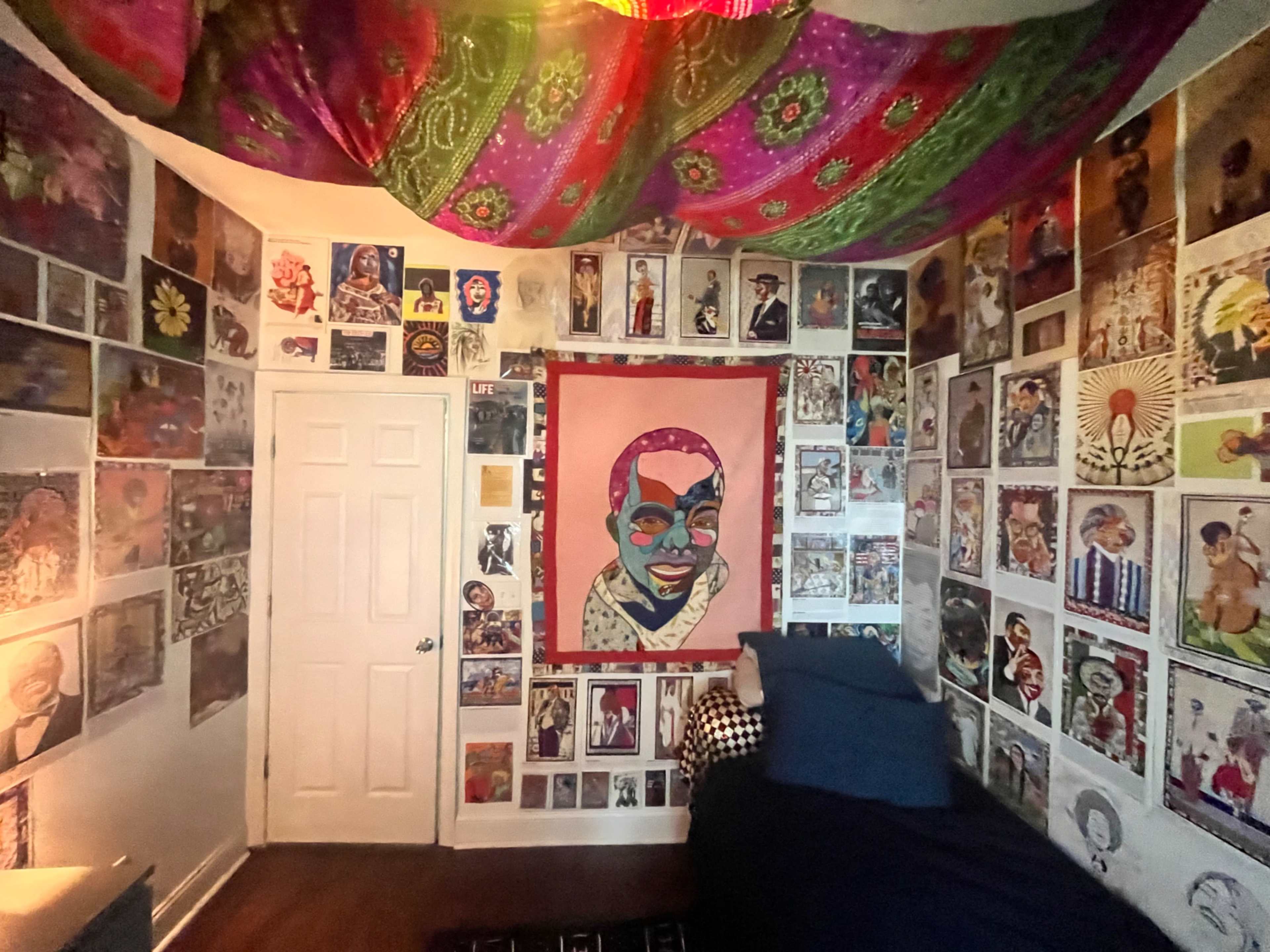 The image shows a vibrant bedroom filled with colorful artwork on the walls and a patterned fabric draping from the ceiling.