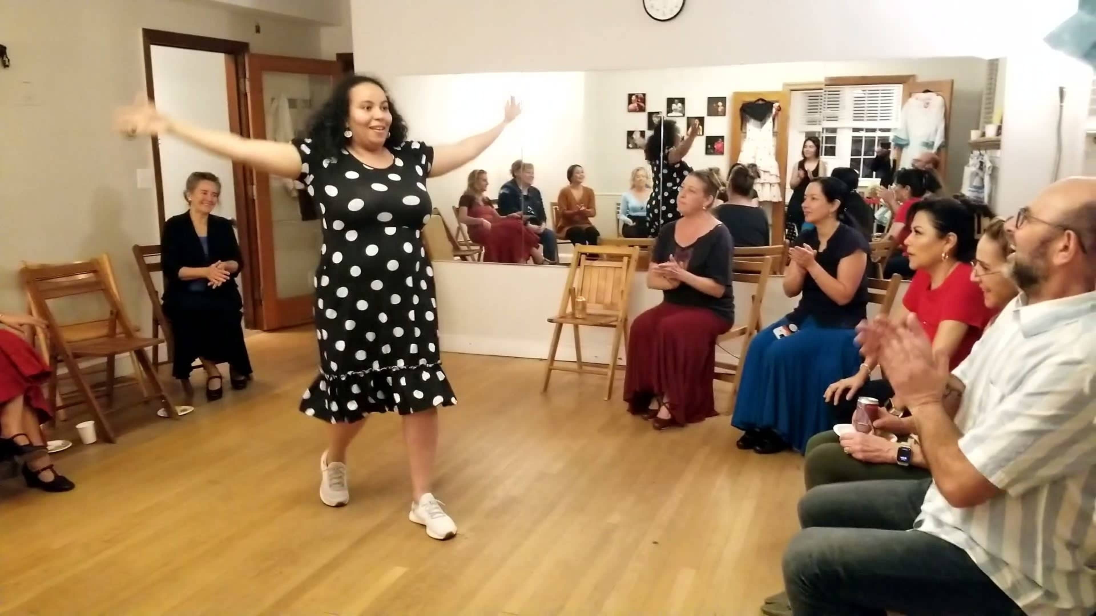 A woman in a polka dot dress performs in front of an audience seated in a dance studio.