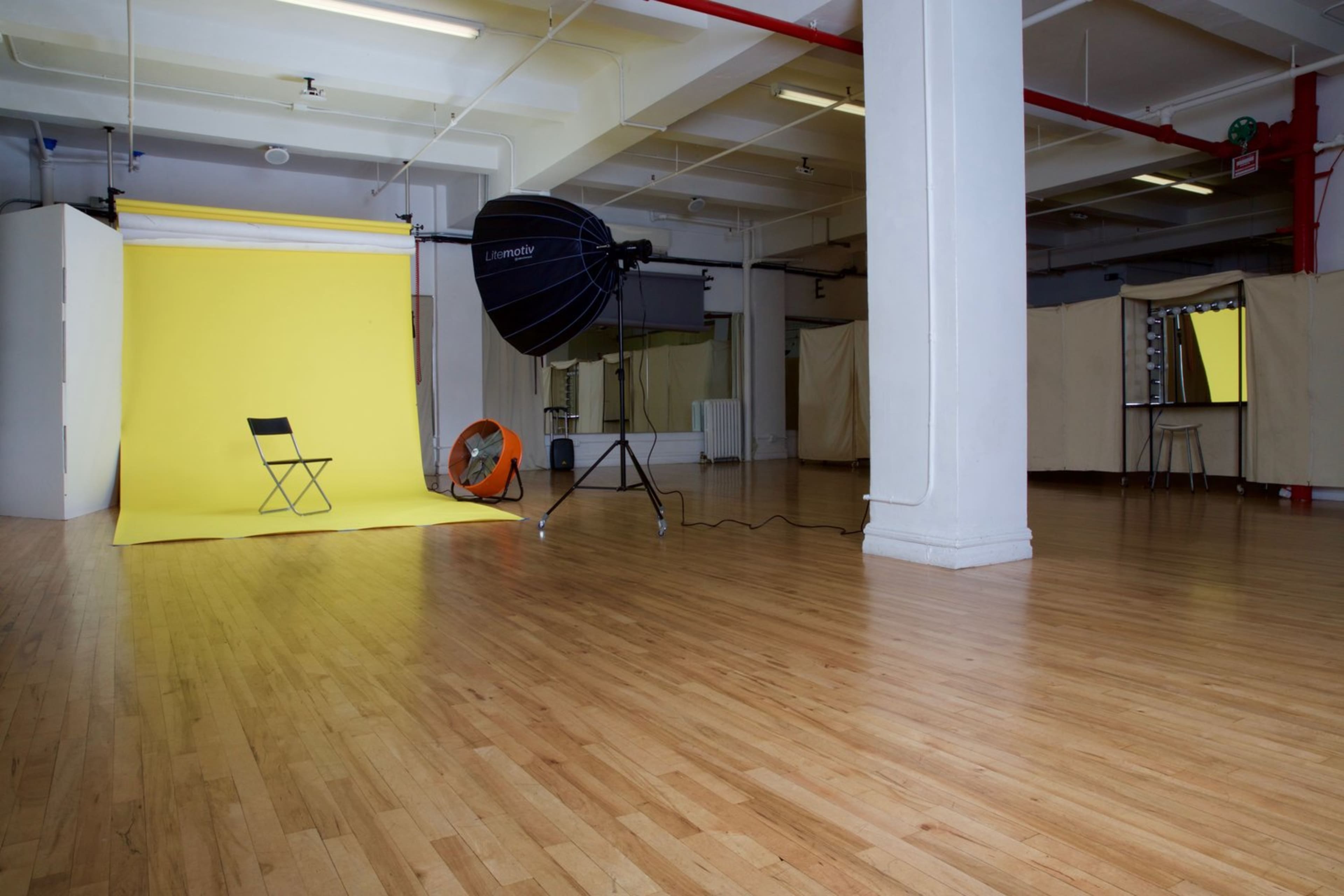 The image shows a photography studio featuring a yellow backdrop, a single chair, a large light source, and a fan on a wooden floor.