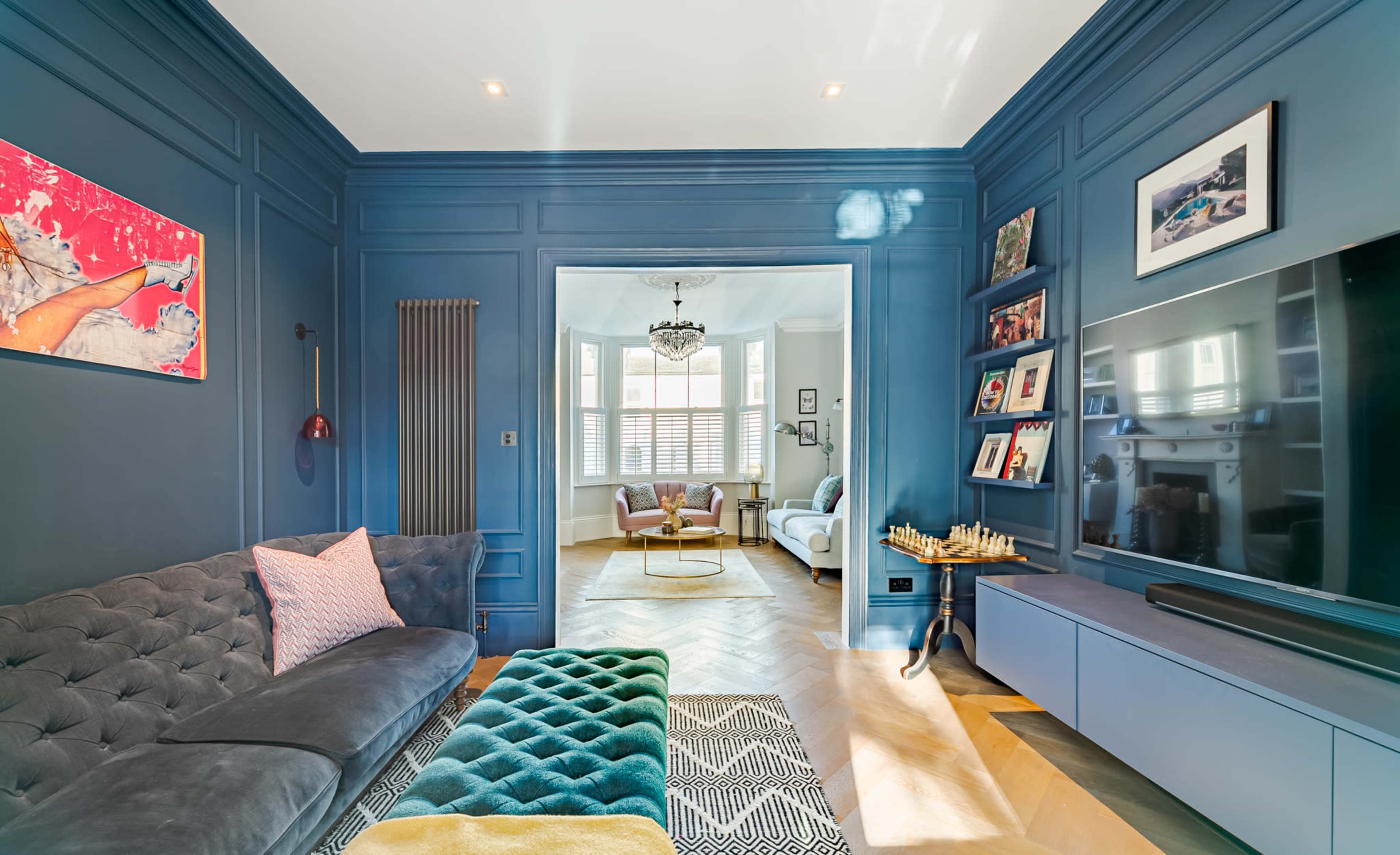 The image shows a living room with a dark blue wall, a gray sofa, and a view into a bright, adjacent room featuring large windows and another seating area.