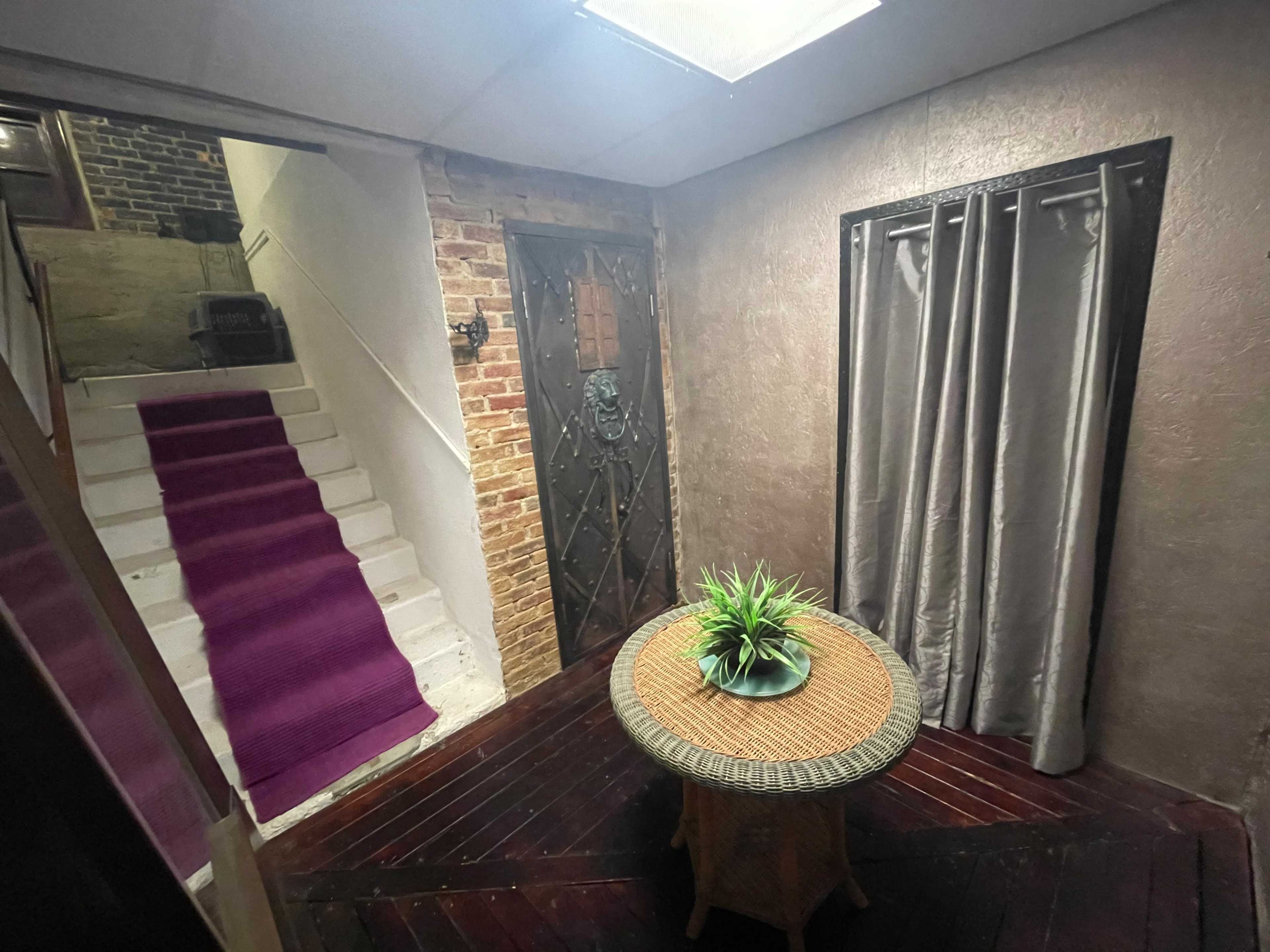 An entrance hall featuring a round table with a plant, a large door with a lion's head knocker, and a staircase with a purple runner leading to an upper level.