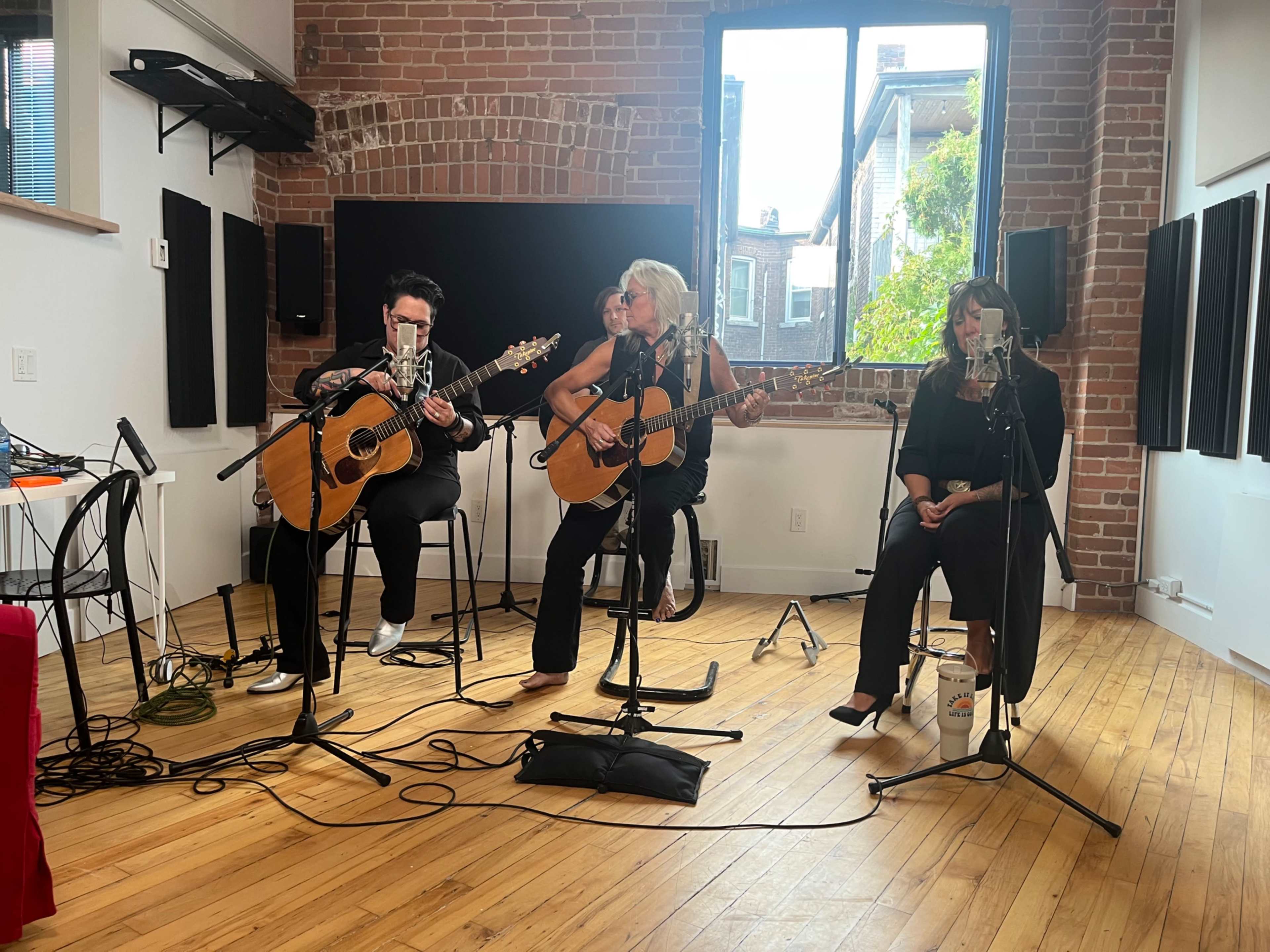 The image shows four musicians performing acoustically in a studio with brick walls and large windows letting in natural light.
