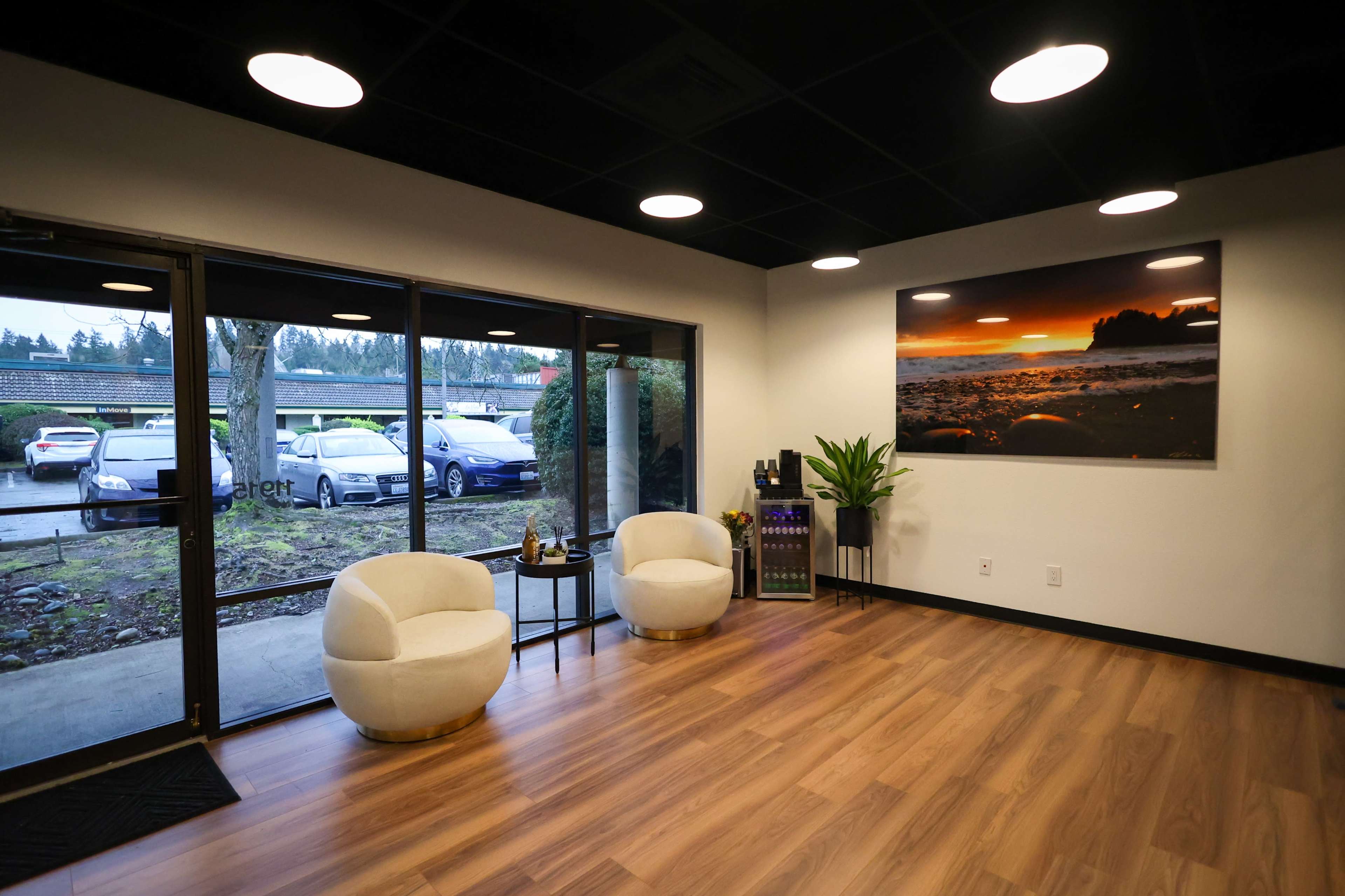 A modern lobby area with two white chairs, a large framed photograph on the wall, and a glass door leading to an outside parking lot.