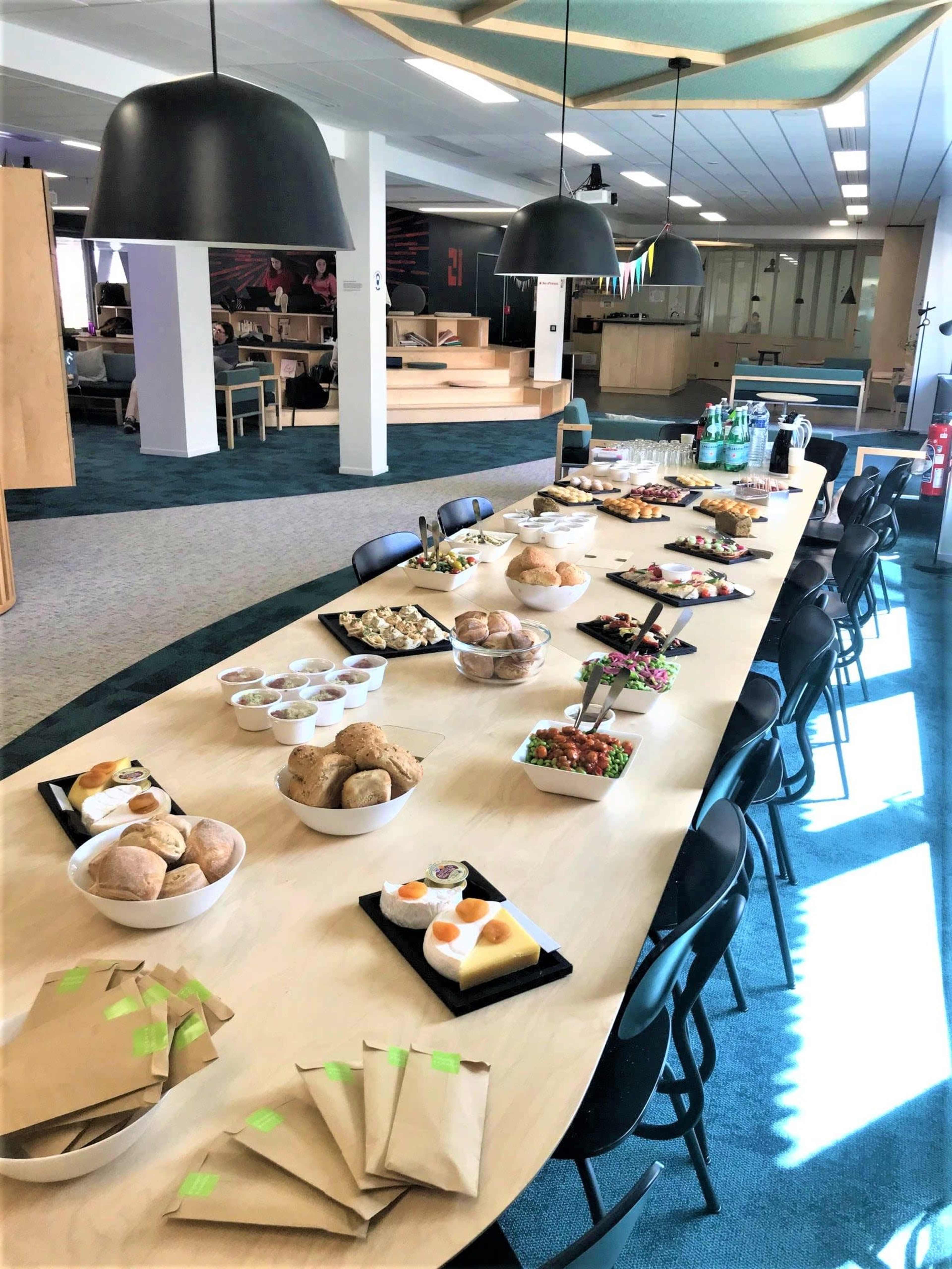 A long table is set with a variety of food items, including pastries, cakes, and salads, in a well-lit room with modern decor.
