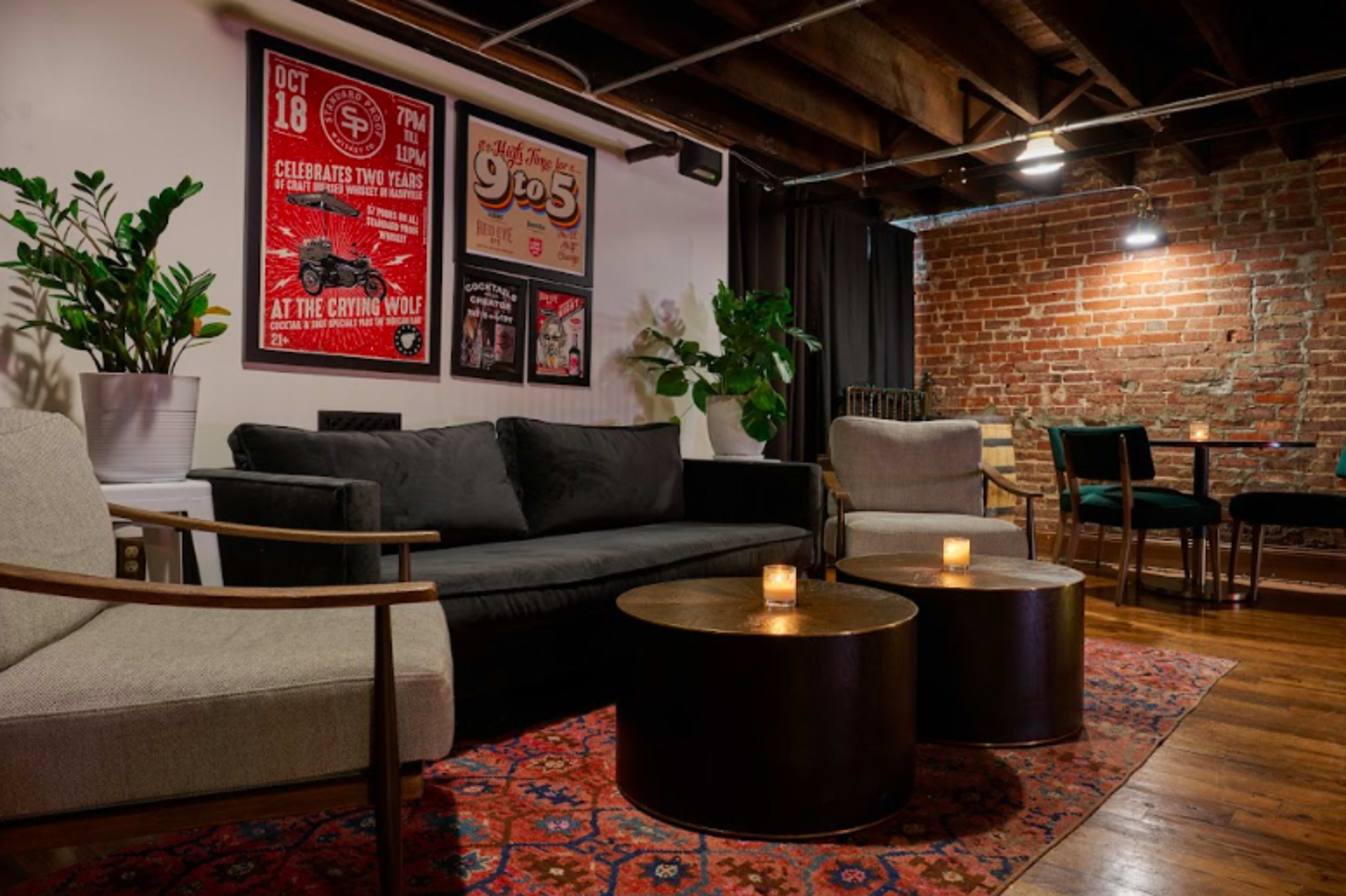 The image shows a cozy lounge area with exposed brick walls, a black sofa, two armchairs, and a pair of small round tables with candles.