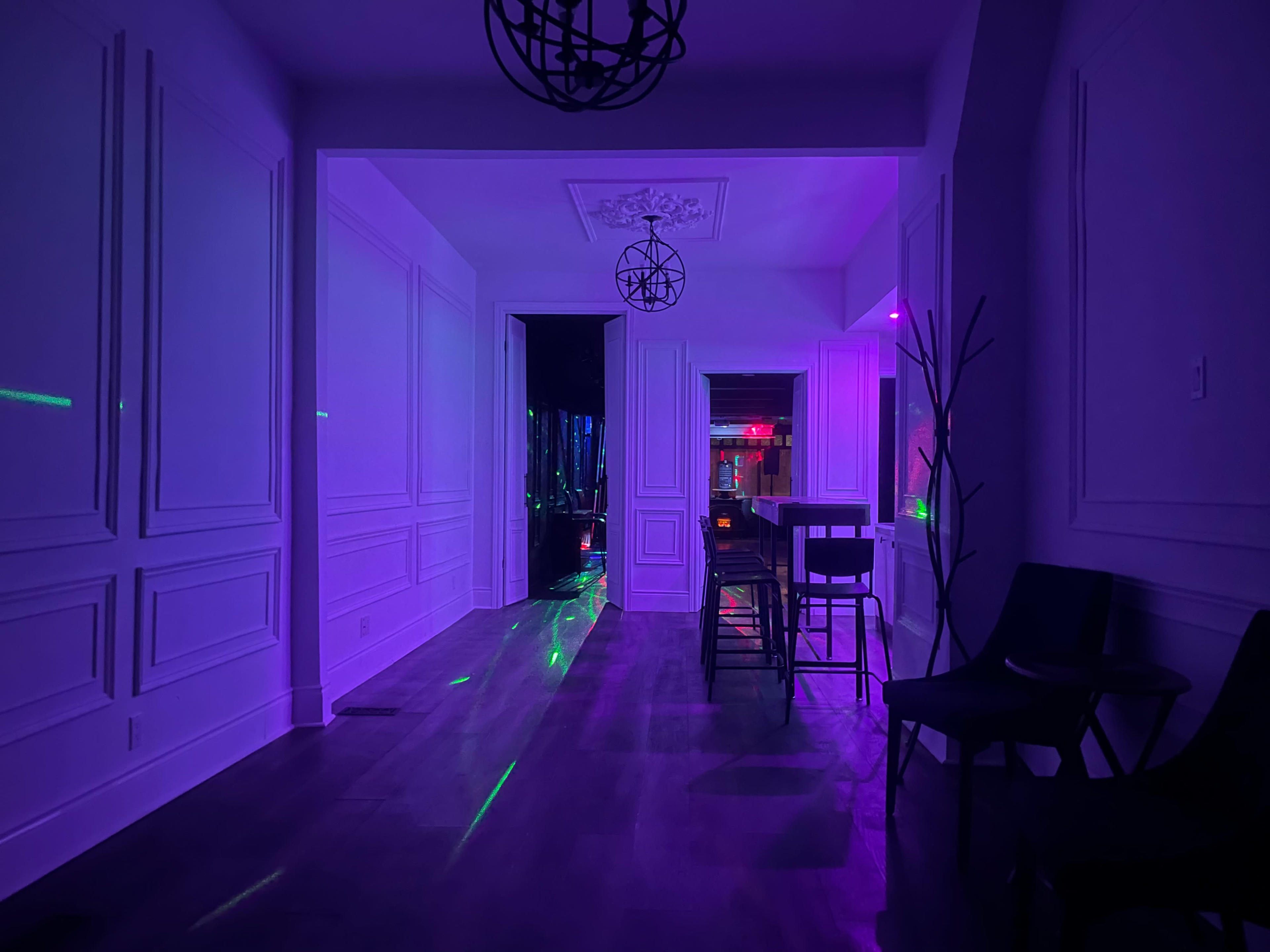 A dimly lit hallway with purple lighting, leading to a room illuminated by colorful lights and featuring bar stools and a wooden table.