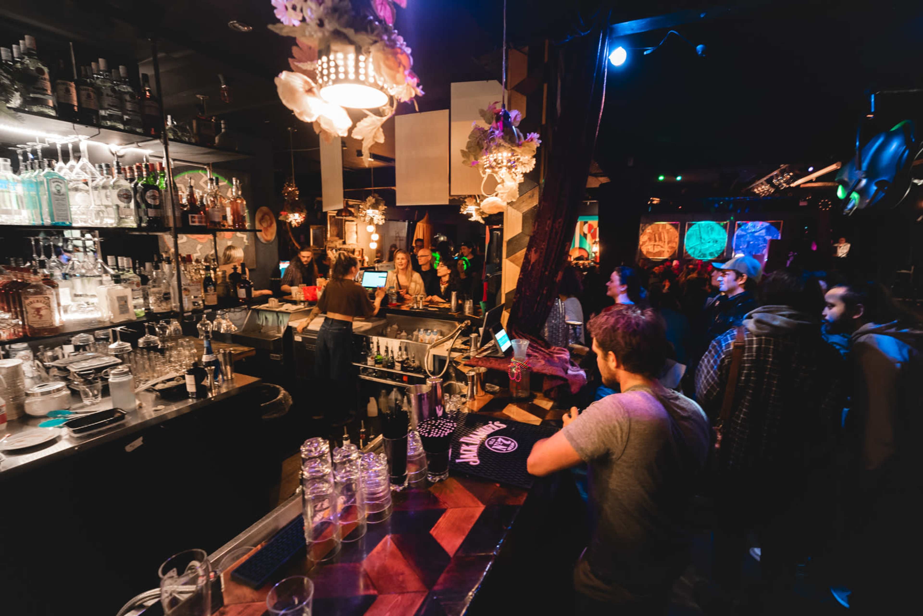 A dimly lit bar scene shows patrons socializing near a well-stocked bar with illuminated bottles and decorative elements.