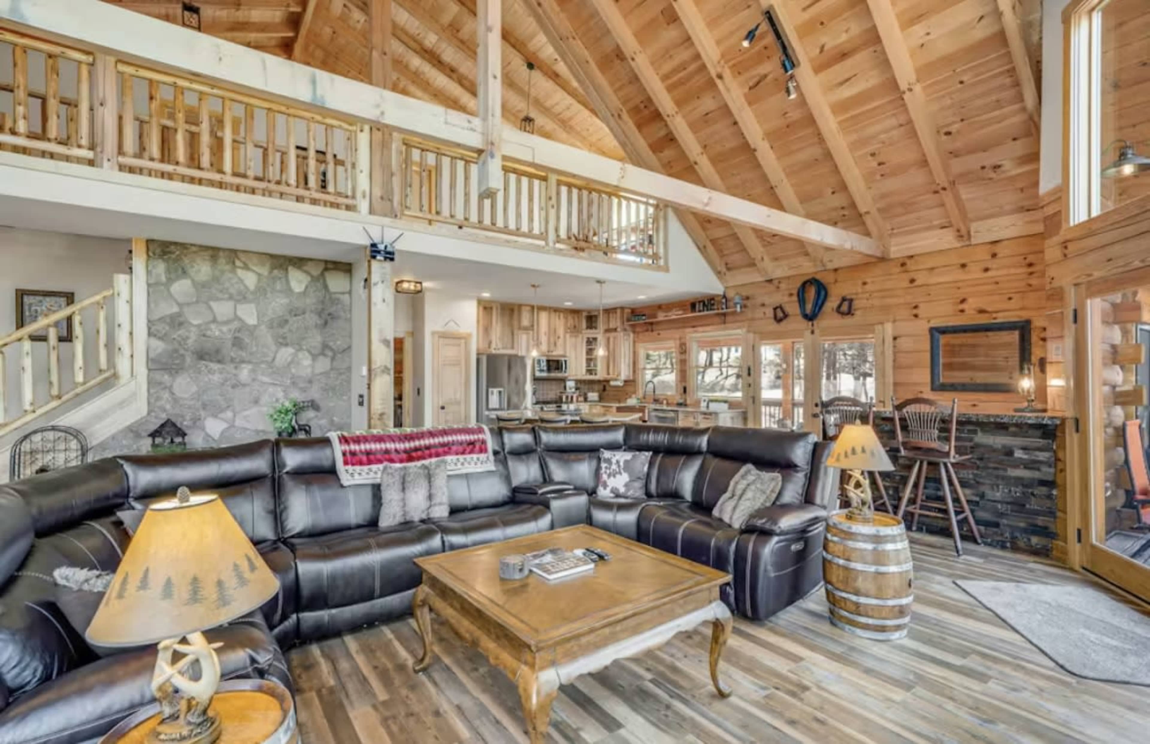 The image depicts a cozy, wooden cabin interior featuring a large leather sectional sofa, wooden beams, and a stone fireplace, with an open floor plan leading to a kitchen area.