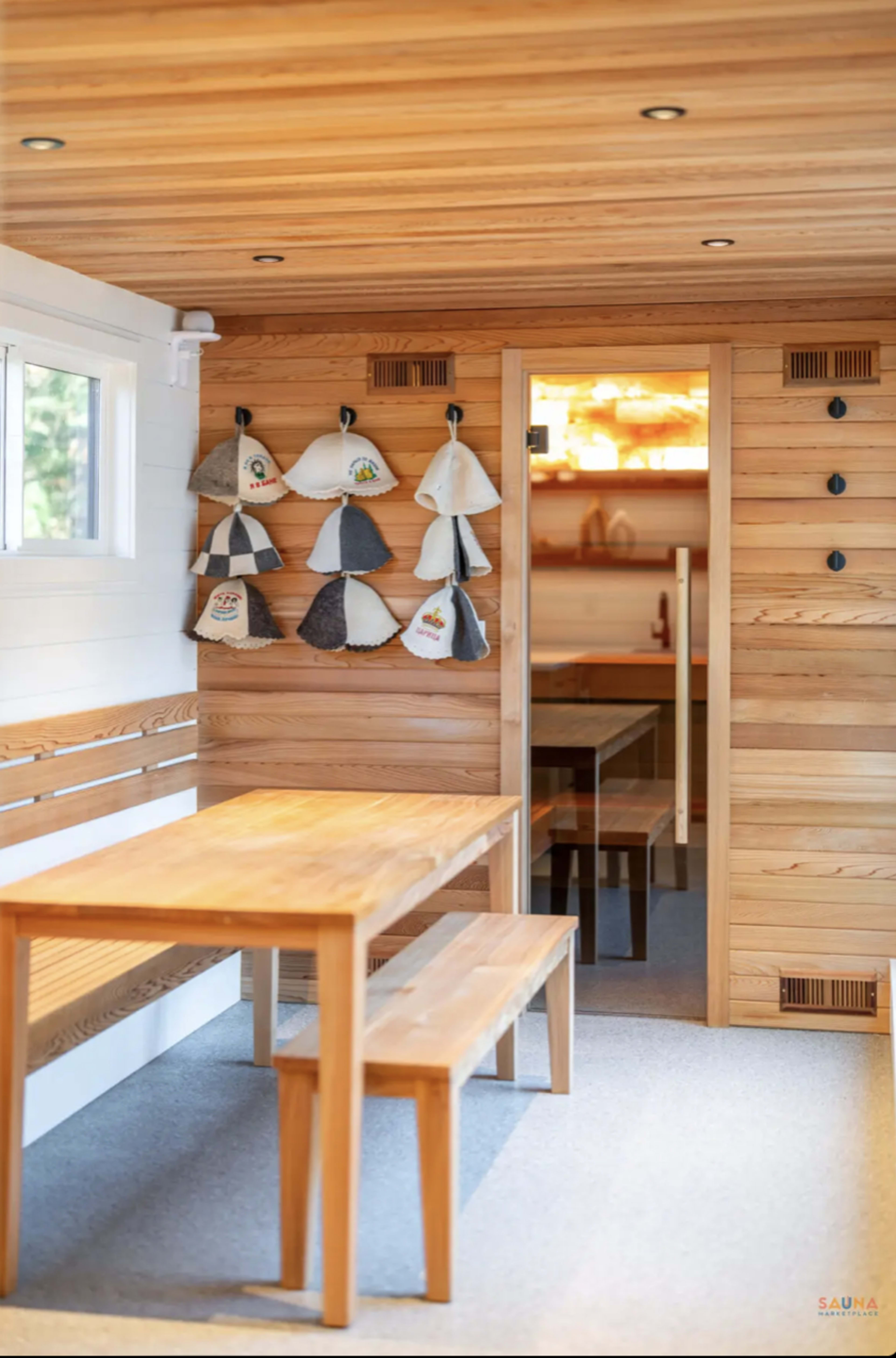 The image shows a wooden sauna interior with a table, benches, and hats hanging on the wall.