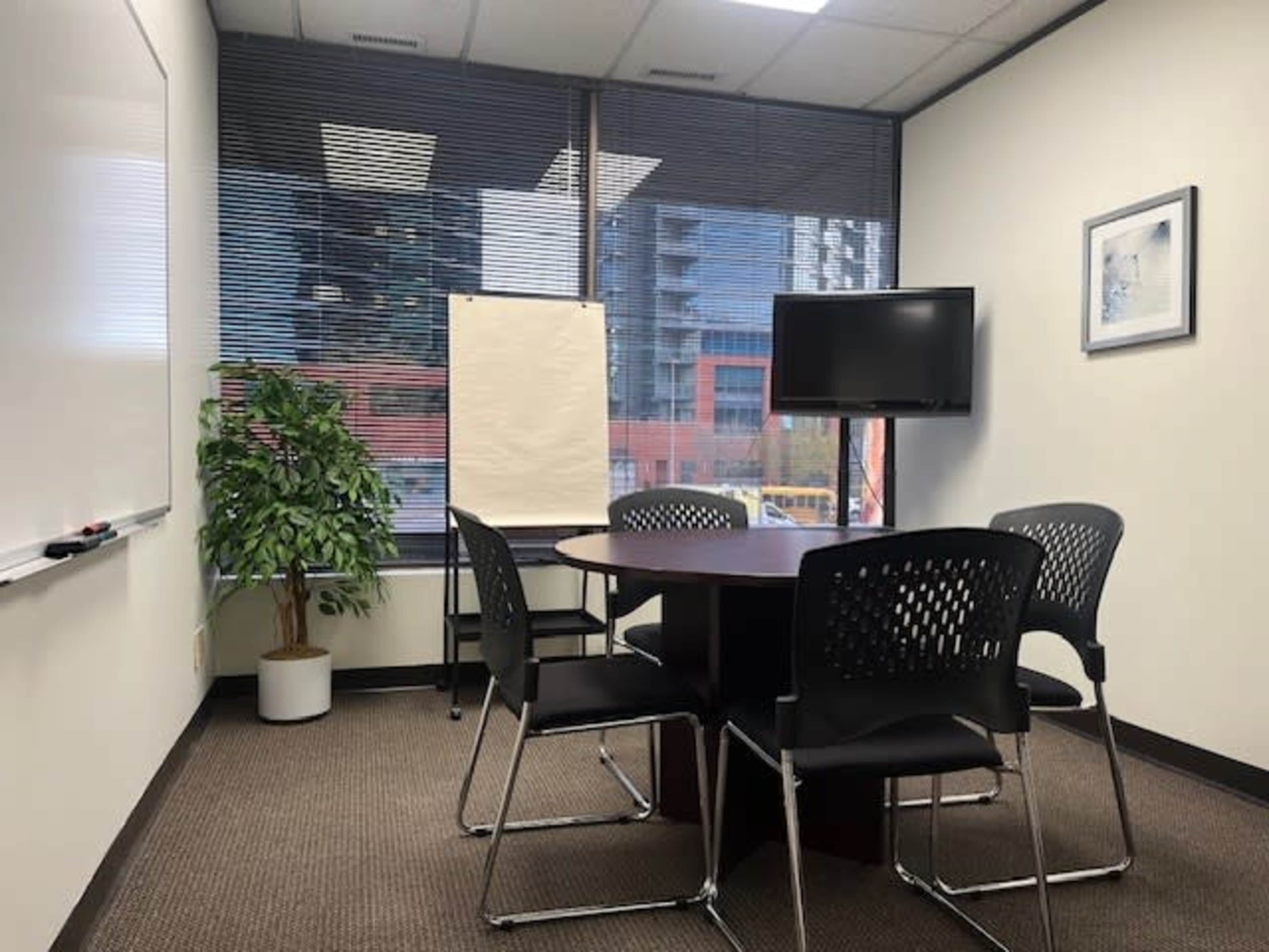 The image shows a meeting room with a round table, four black chairs, a whiteboard, a presentation easel, a television monitor, and a potted plant.