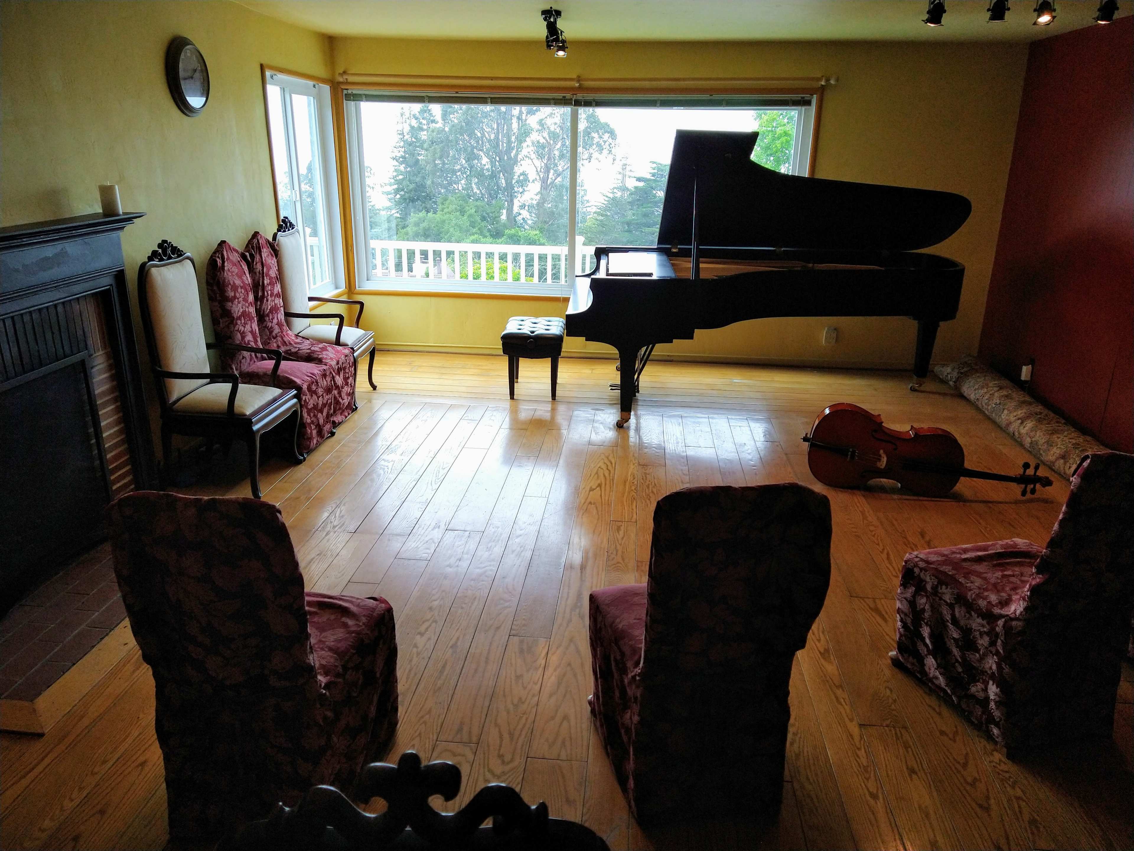 A grand piano is positioned near a large window in a room with wooden floors and four chairs arranged around it.